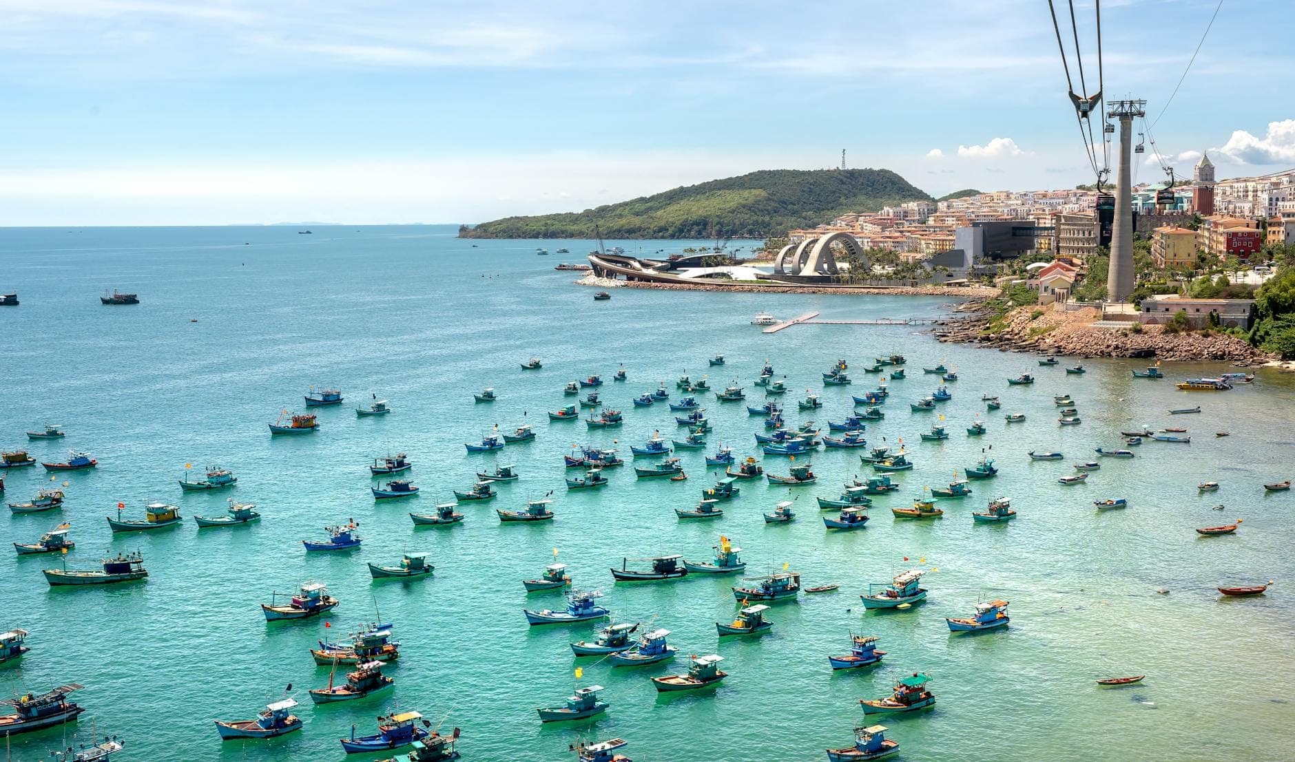 Những chiếc thuyền đánh cá nhiều màu sắc neo đậu trên làn nước xanh ngọc gần An Thới, với tháp cáp treo và các công trình ven biển của đảo Phú Quốc hiện ra phía sau.