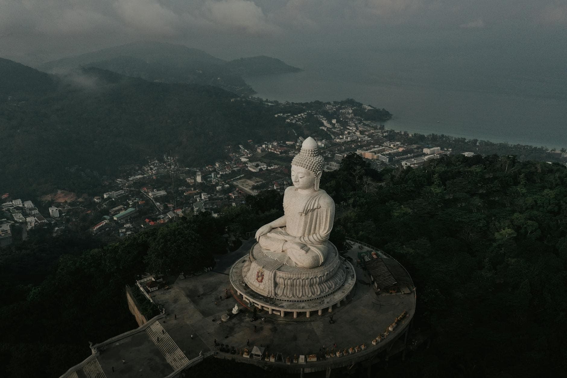 Aerial view of the Big Buddha statue overlooking Phuket island, lush green hills, and coastline under a misty sky.