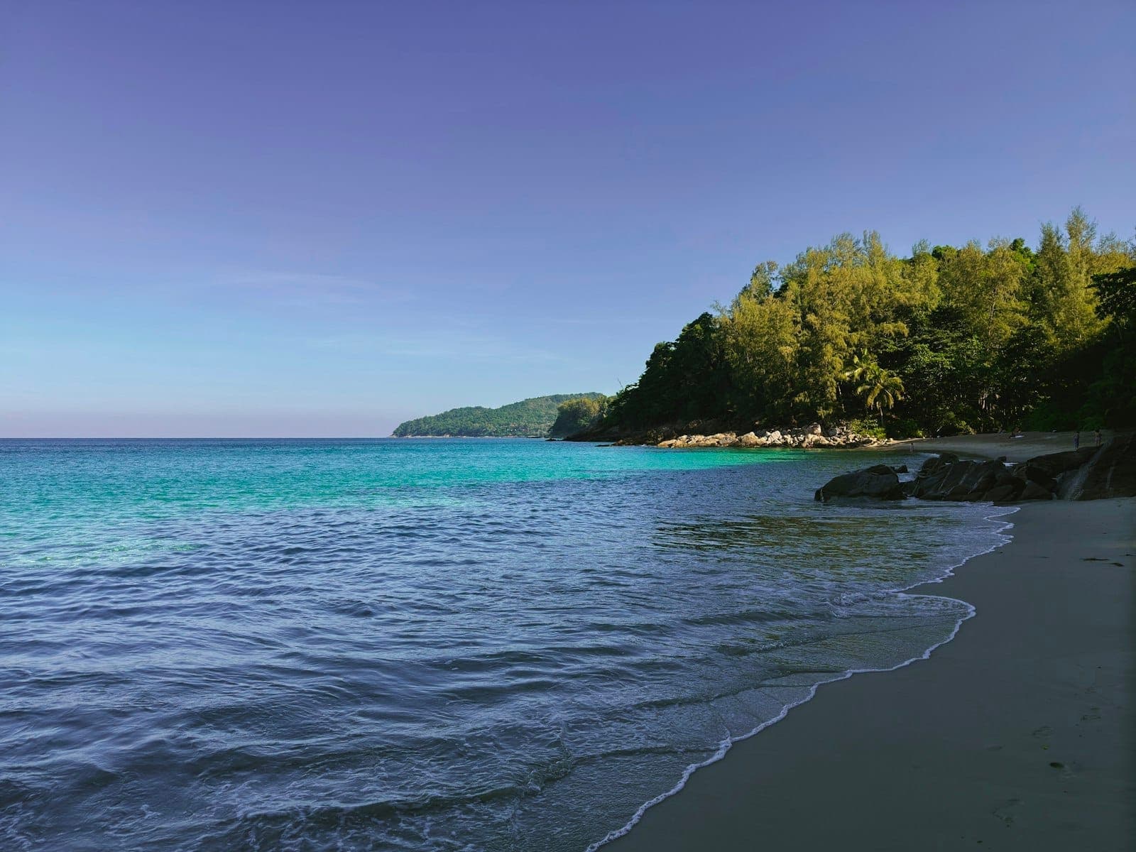 Malawak at walang taong dalampasigan ng Phuket na may turquoise na tubig, kagubatang tangway, at walang tao, na nagpapahiwatig ng kalawakan at katahimikan.