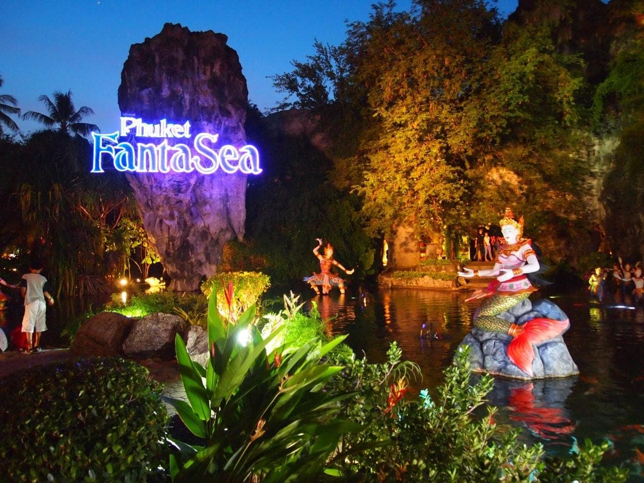Brightly lit Phuket FantaSea sign beside a garden pond with colorful mermaid statue and lush greenery at twilight.