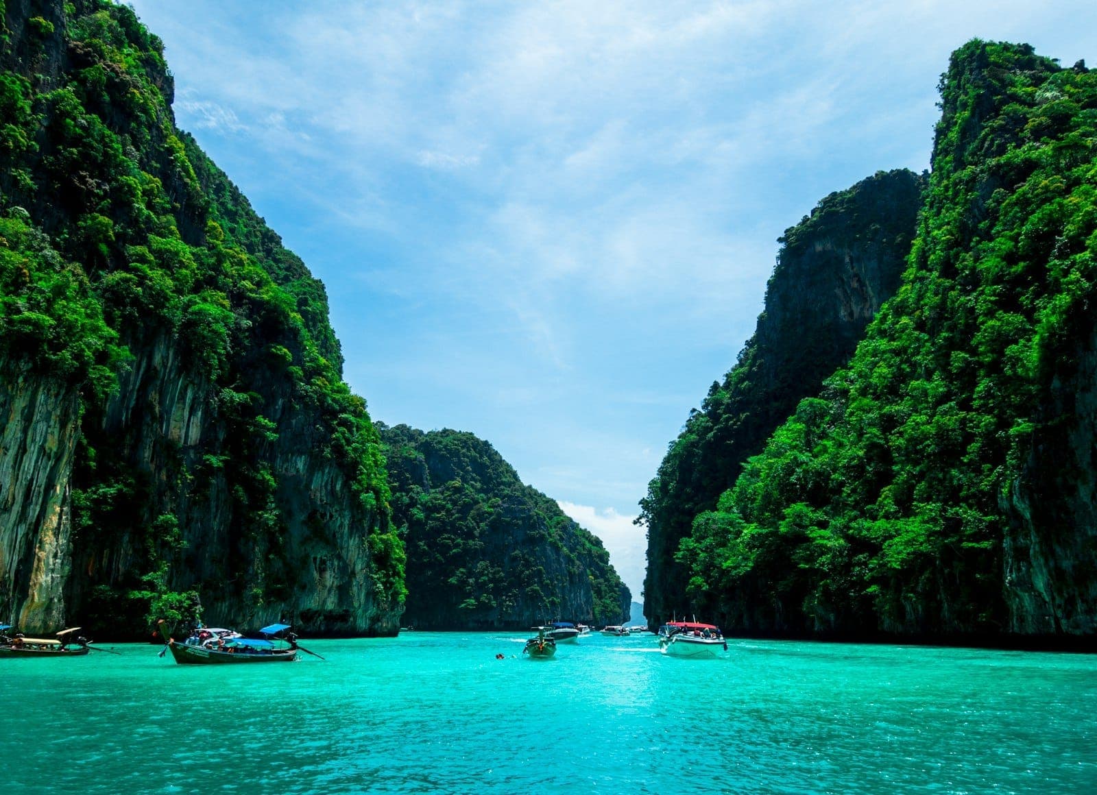 Turquoise water surrounded by dramatic limestone cliffs, with several longtail boats and speedboats enjoying the scenic bay under a blue sky.