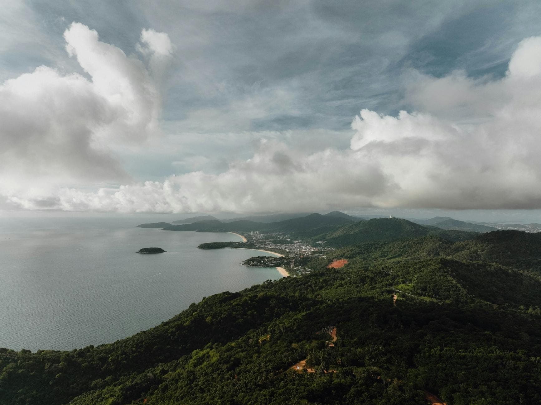 Large vue aérienne du littoral de Phuket avec ses collines verdoyantes, son rivage, la mer et des nuages épars au-dessus.