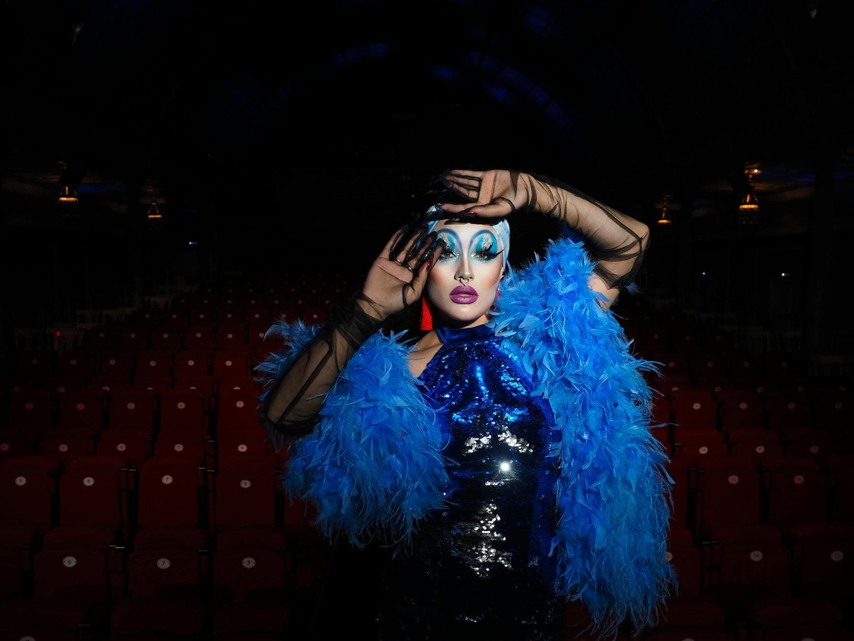 นักแสดงคาบาเรต์ที่แต่งกายฉูดฉาดในชุดขนนกสีฟ้าพร้อมเครื่องสำอางเวทีที่โดดเด่น โพสท่าในโรงละครที่ว่างเปล่า