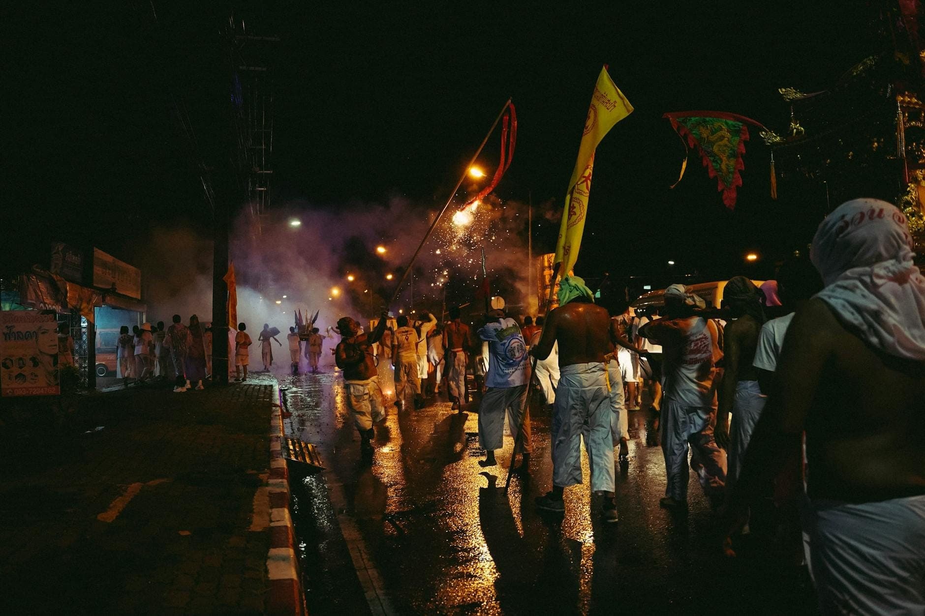 ภาพถนนยามค่ำคืนในภูเก็ตที่เต็มไปด้วยฝูงชนและธงเทศกาล ผู้คนกำลังเฉลิมฉลองในงานที่คึกคักใต้แสงไฟถนนและท้องฟ้าที่มีควัน