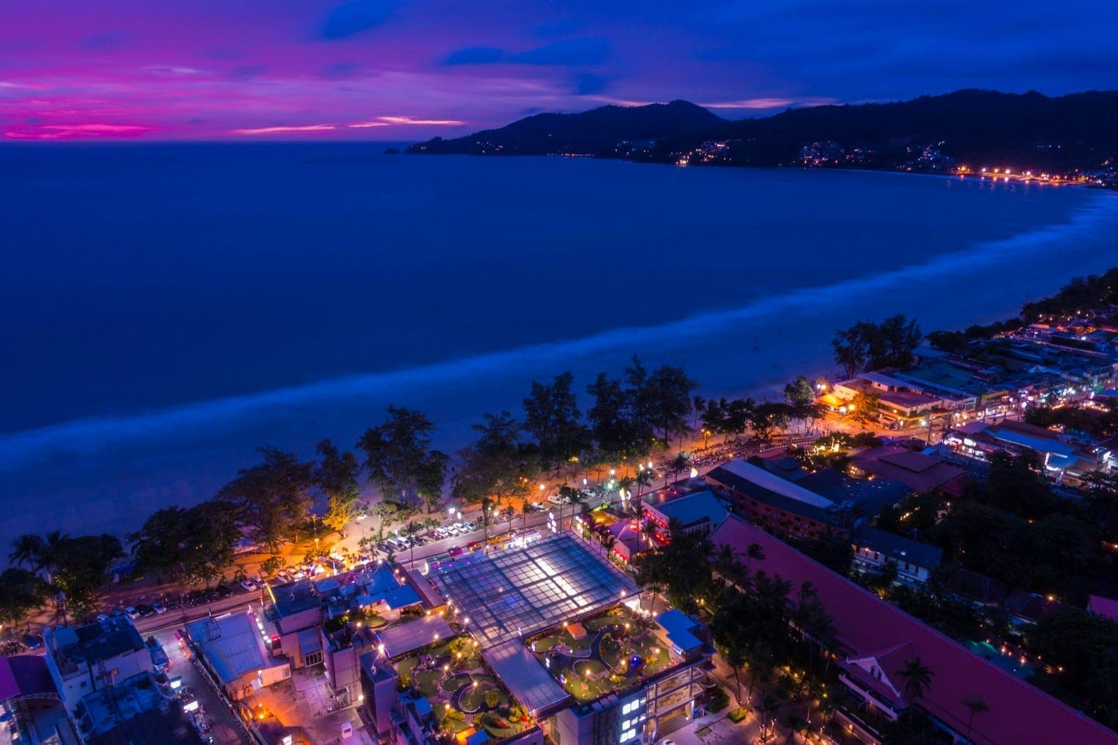 A stunning aerial view of Phuket’s coastline at night, with brightly lit clubs and bars near the beach, glowing against a vibrant purple and blue sky.