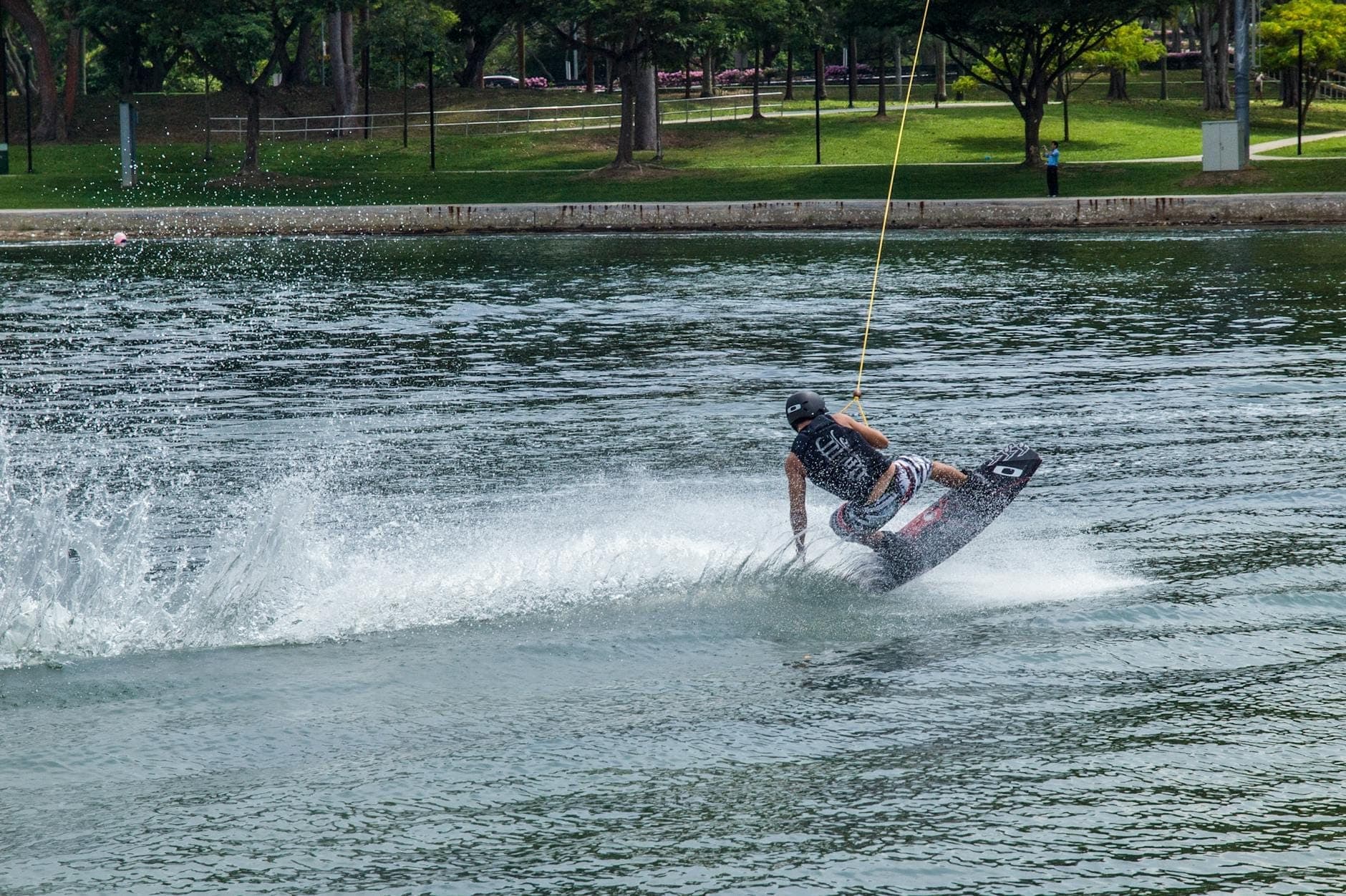 Wakeboarder gliding across the lake at Phuket Wake Park, surrounded by green trees and grassy park under bright daylight.