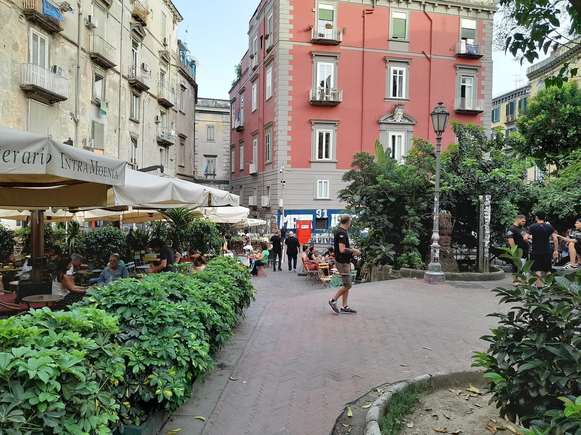 Vista da Piazza Bellini em Nápoles com cafés ao ar livre, vegetação exuberante, edifícios históricos e pessoas aproveitando a animada praça durante o dia.