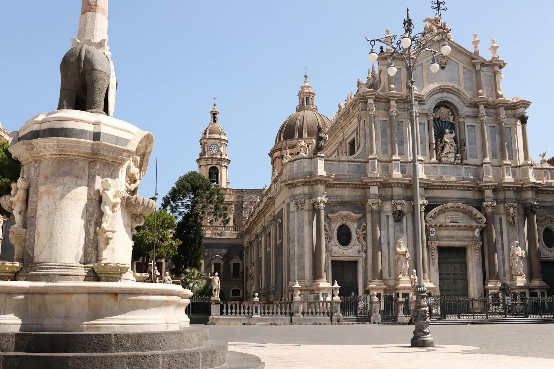 Pemandangan Piazza del Duomo di Catania dengan Fontana dell'Elefante yang ikonik dan Katedral Catania bergaya Barok di latar belakang, di bawah langit cerah dengan detail arsitektur yang memukau.