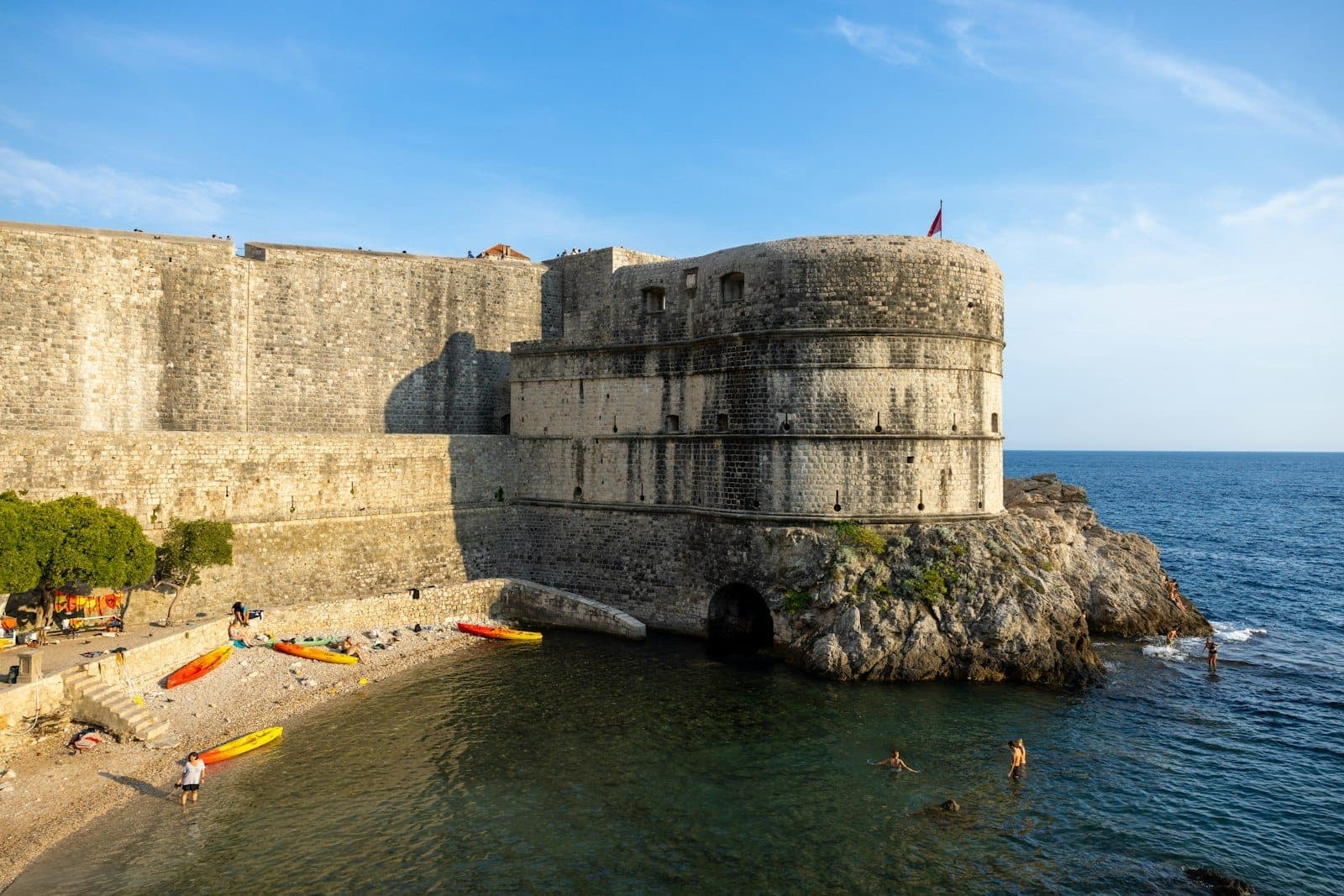 Blick auf die Festungsmauer des Pile-Tors in Dubrovnik, die aus dem Meer aufsteigt – mit Menschen am Strand, Kajaks und strahlend blauem Himmel.