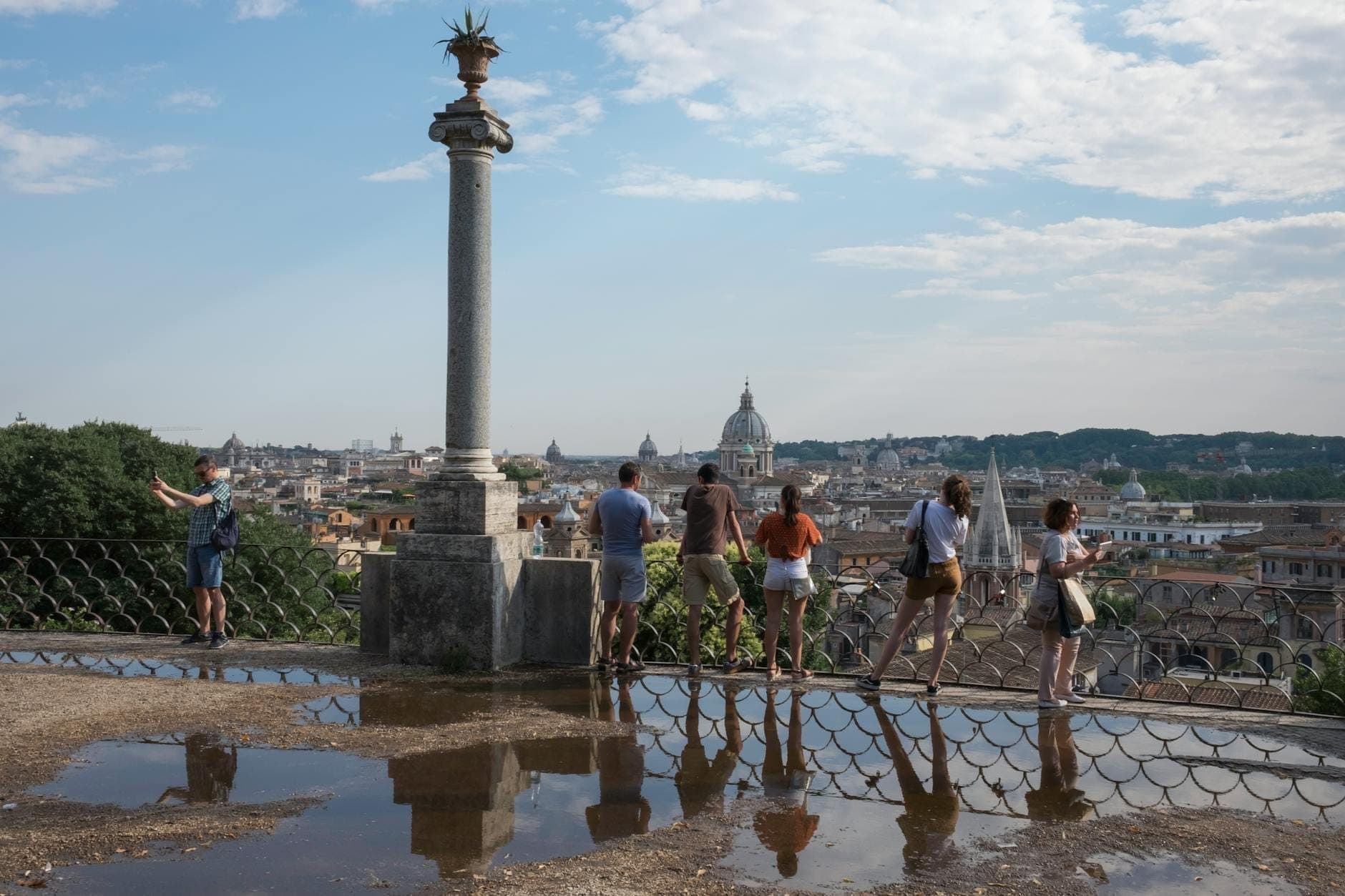 Mga bisita na nag-eenjoy ng panoramikong tanawin mula sa Pincio Terrace sa ibabaw ng mga bubungan ng Roma, na may nakikitang domo ng Basilica ng San Pedro sa malayo sa ilalim ng asul na langit.