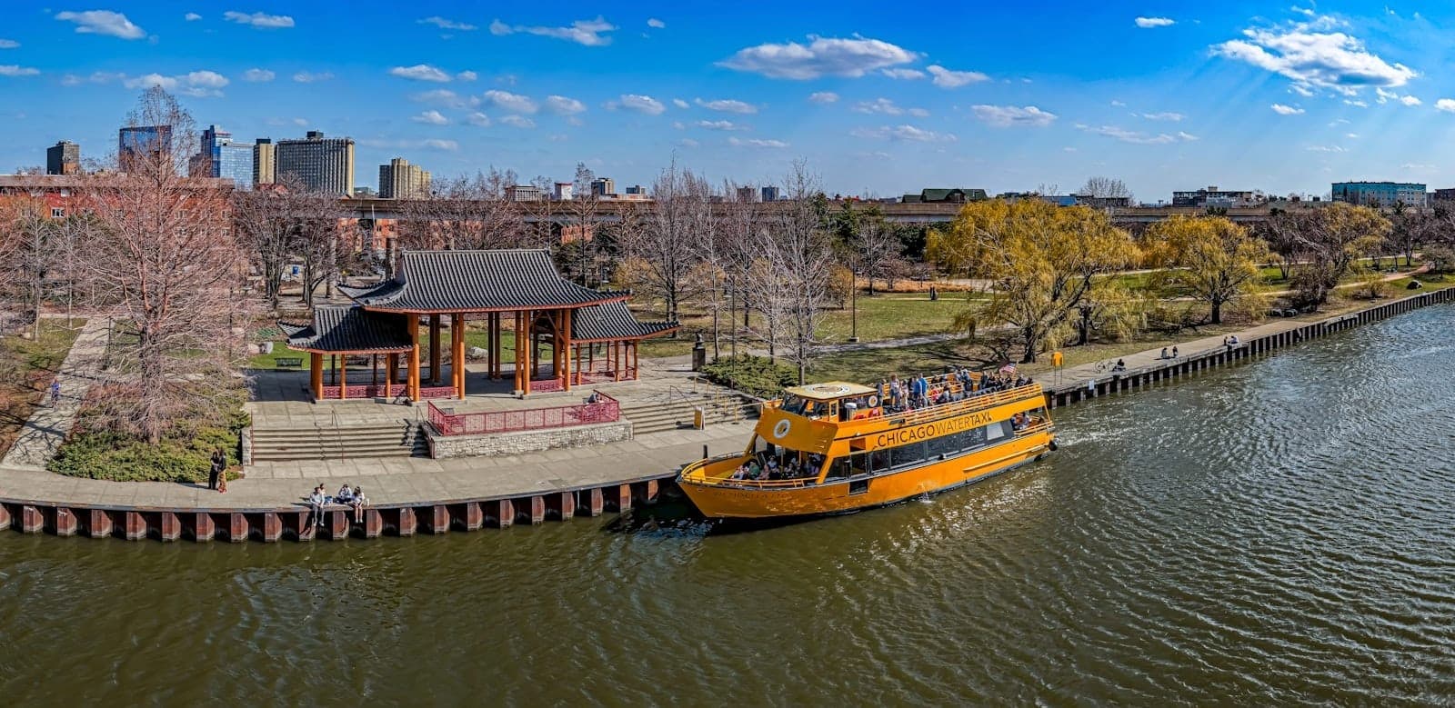 A bright panoramic view of Ping Tom Memorial Park’s riverfront pavilion with a yellow water taxi docked along the Chicago River on a clear sunny day.