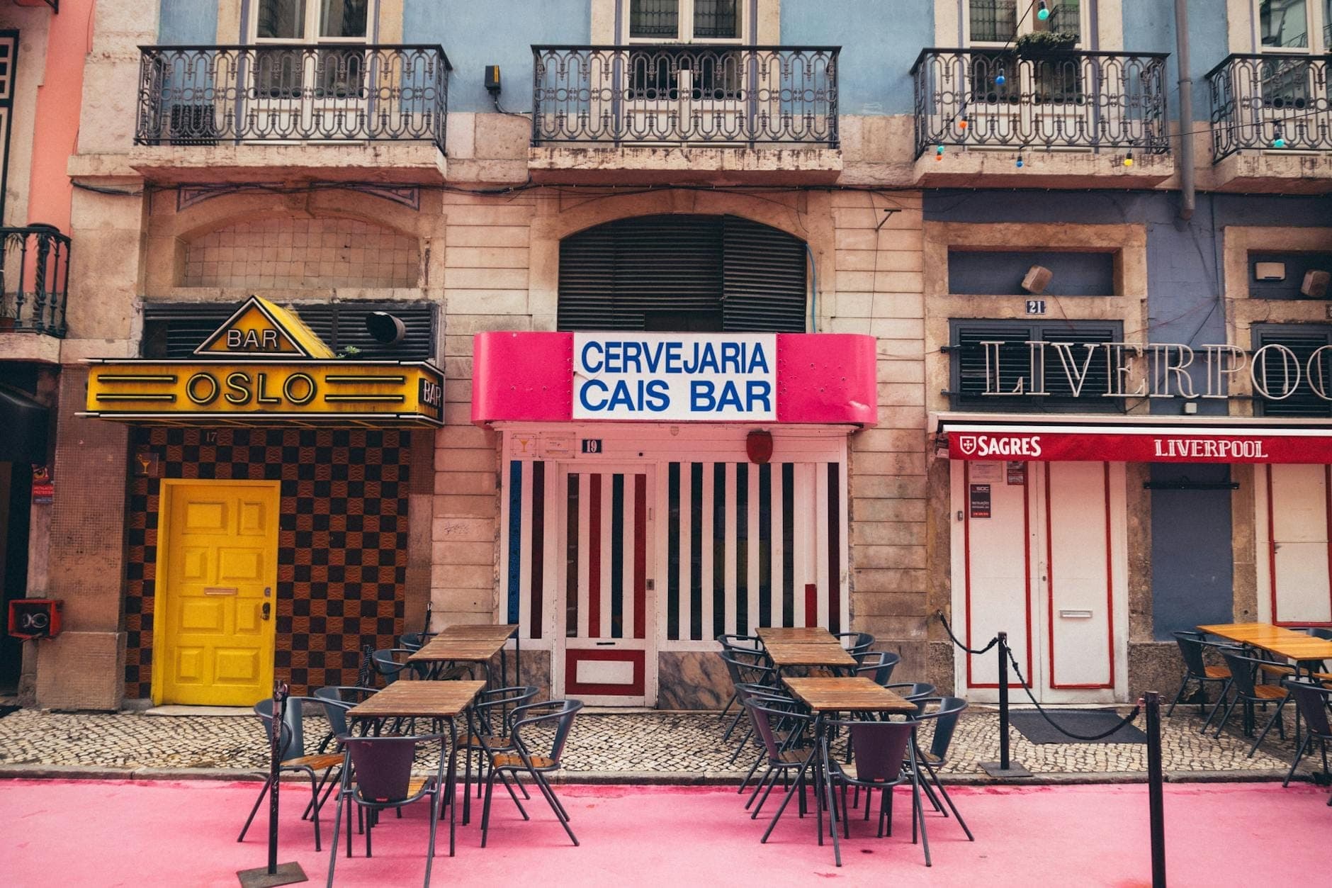 Fachadas vintage coloridas com mesas do lado de fora alinhadas ao longo do calçadão rosa da Rua Nova do Carvalho, a famosa Pink Street de Lisboa, durante o dia.
