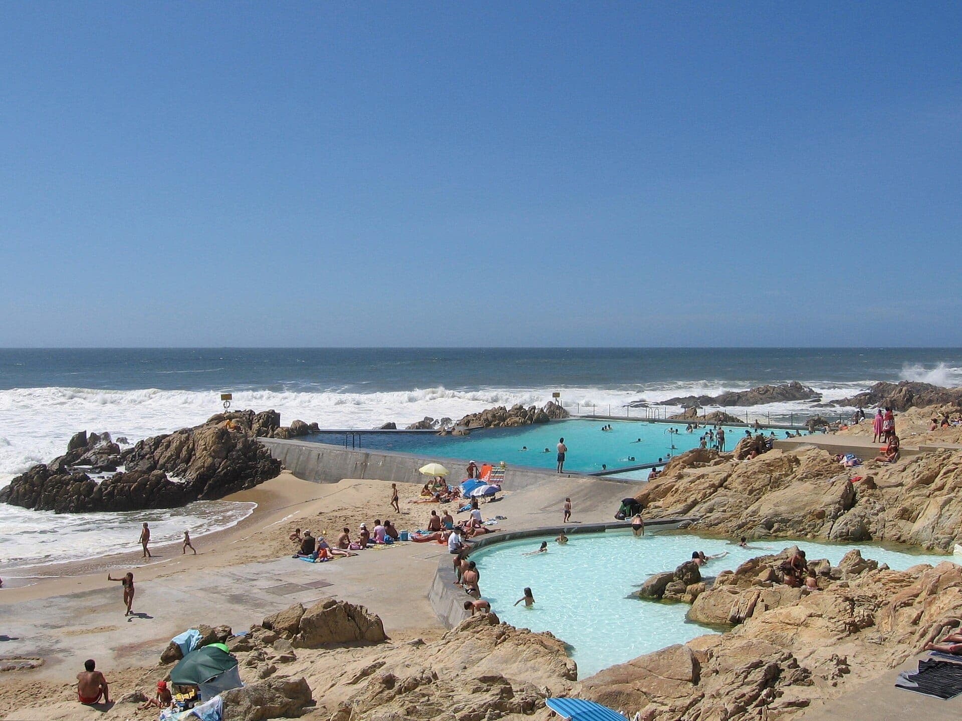 Die Schwimmbecken der Piscina das Marés fügen sich nahtlos in die felsige Atlantikküste ein, mit Sonnenbadenden, Schwimmern und Naturstein unter einem klaren blauen Himmel.