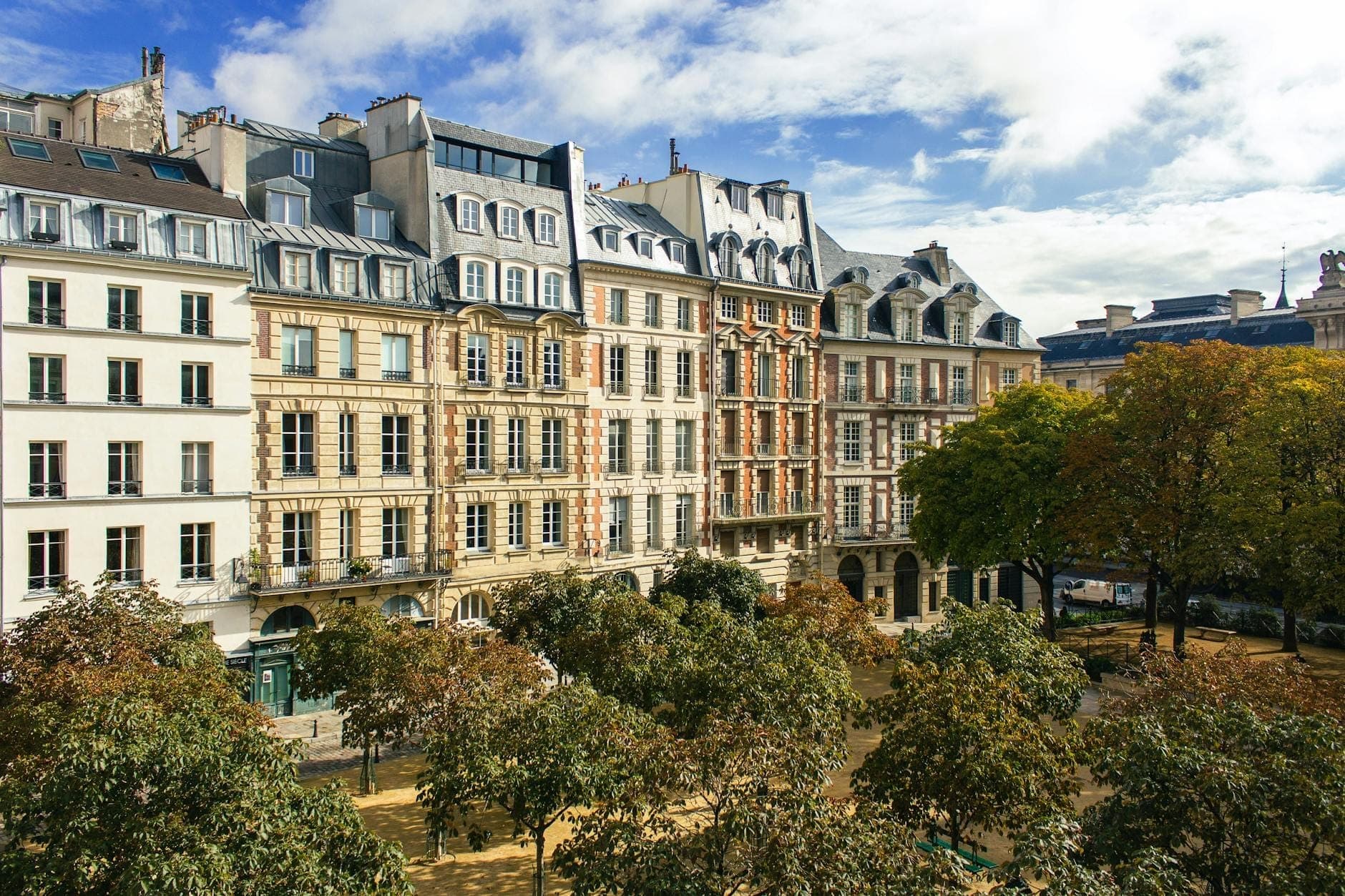 Elevated view of Place Dauphine with leafy plane trees, classic 17th-century Parisian buildings, and blue sky, capturing the calm charm of the square.