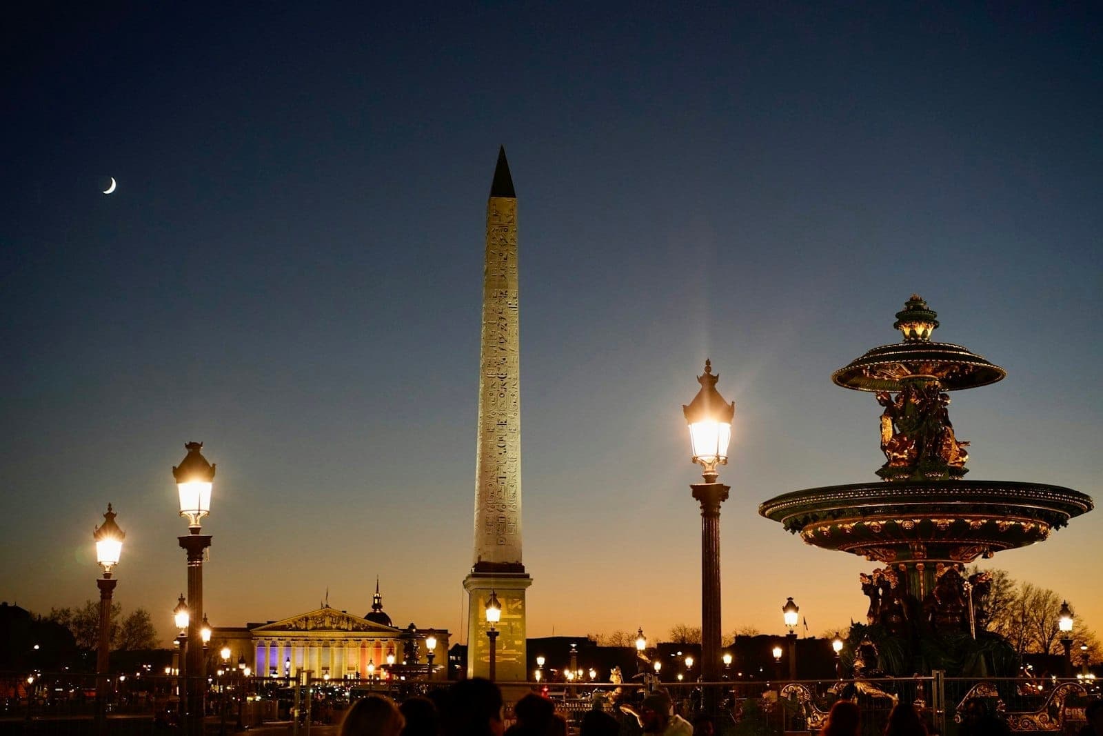 Place de la Concorde at dusk with illuminated street lamps, the Egyptian obelisk, ornate fountain, and people enjoying the lively atmosphere against a twilight sky.
