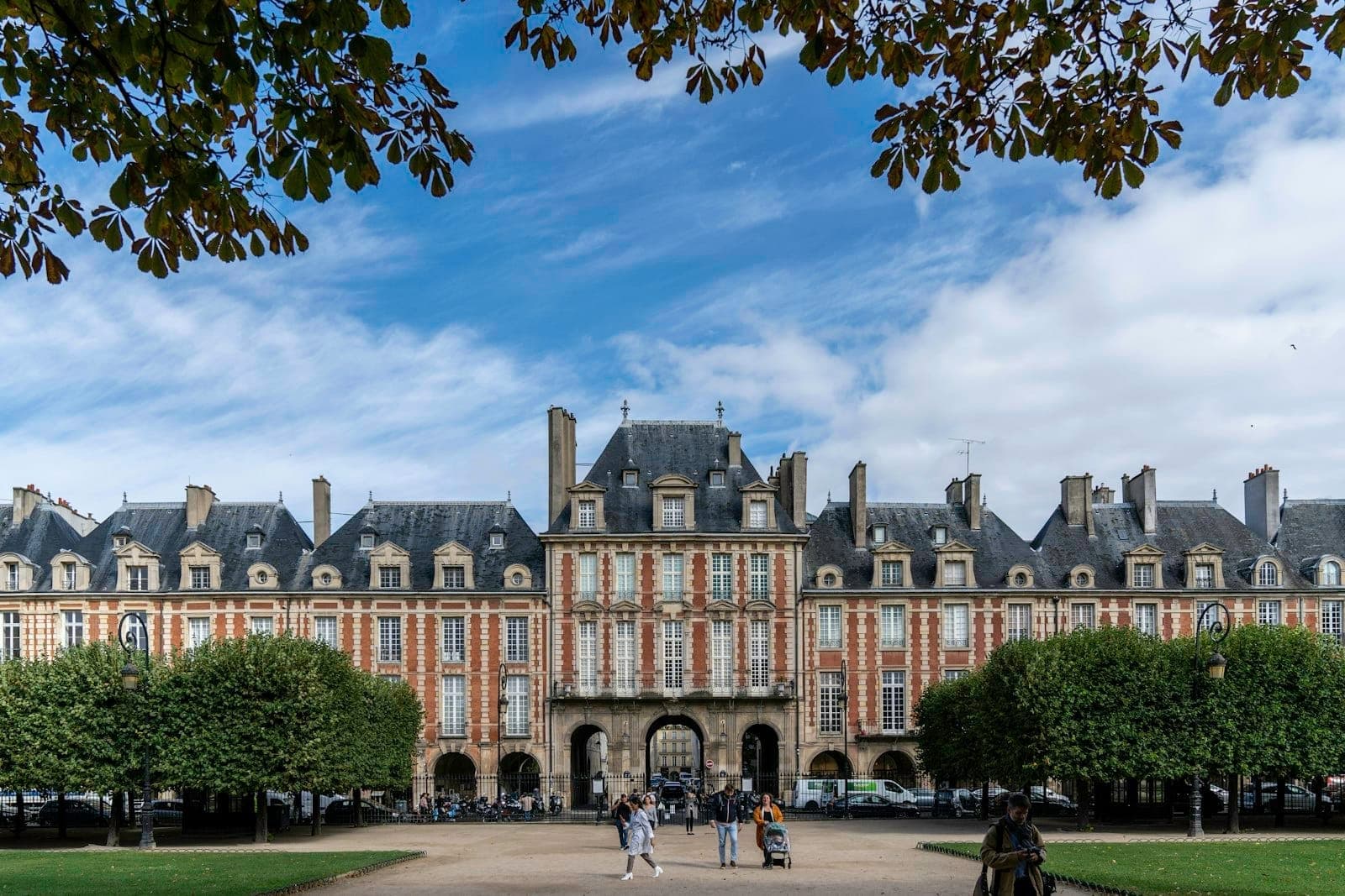 Wide view of Place des Vosges with its red-brick pavilions, formal garden, trees, and people walking under a bright blue sky with clouds.