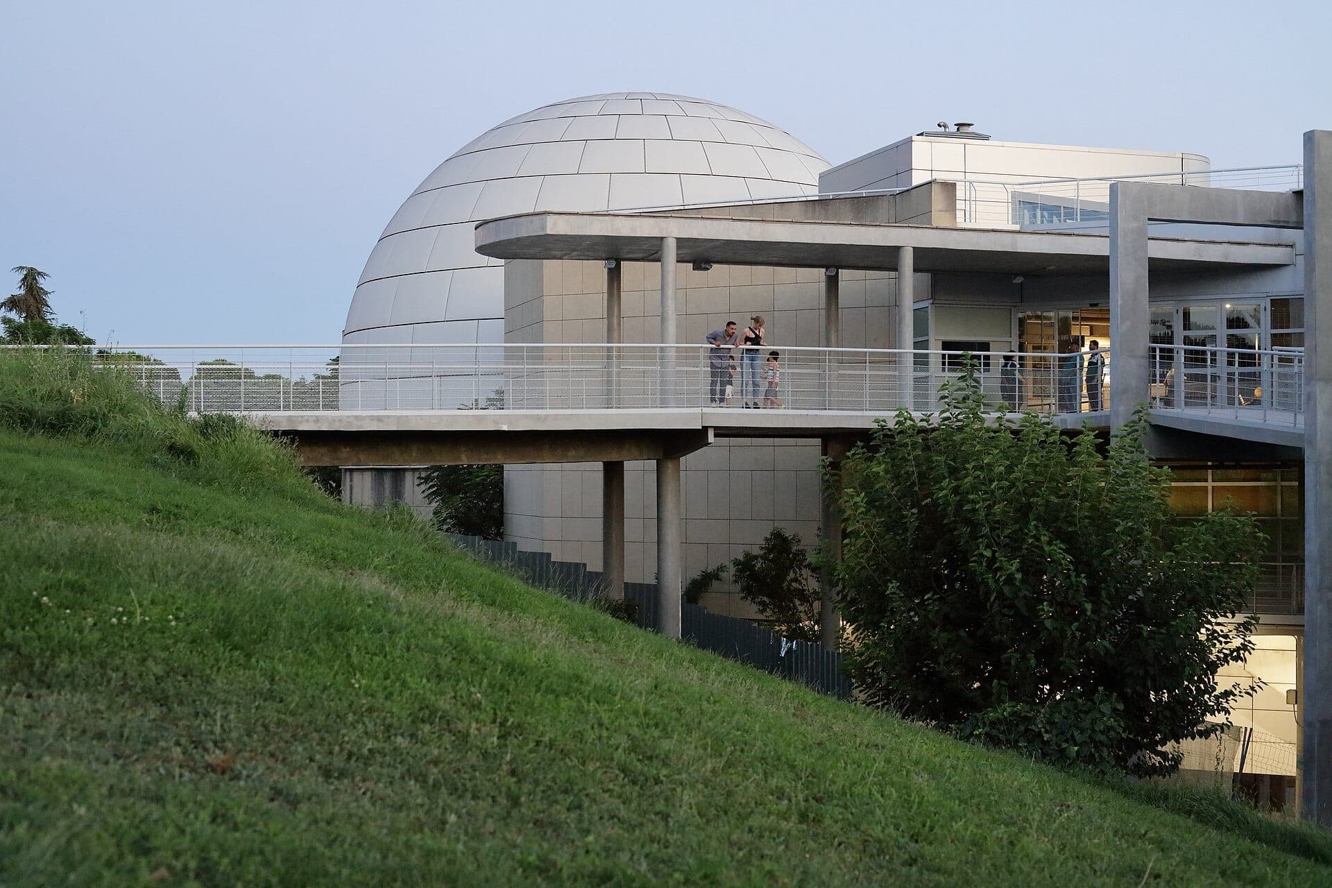 ภายนอกอาคารสมัยใหม่ของ Planetario de Madrid พร้อมโดมอันโดดเด่น เนินหญ้าสีเขียวในเบื้องหน้า และนักท่องเที่ยวที่กำลังเพลิดเพลินยามเย็น