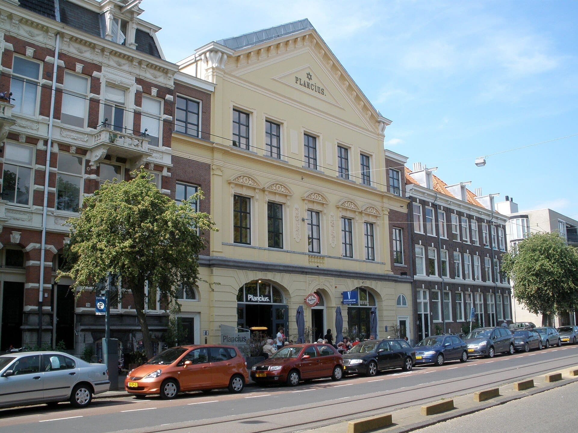 Das historische Plancius-Gebäude mit verzierter gelber Fassade und angrenzenden klassischen niederländischen Stadthäusern im Plantage- und Jüdischen Viertel, Amsterdam, unter einem klaren blauen Himmel.