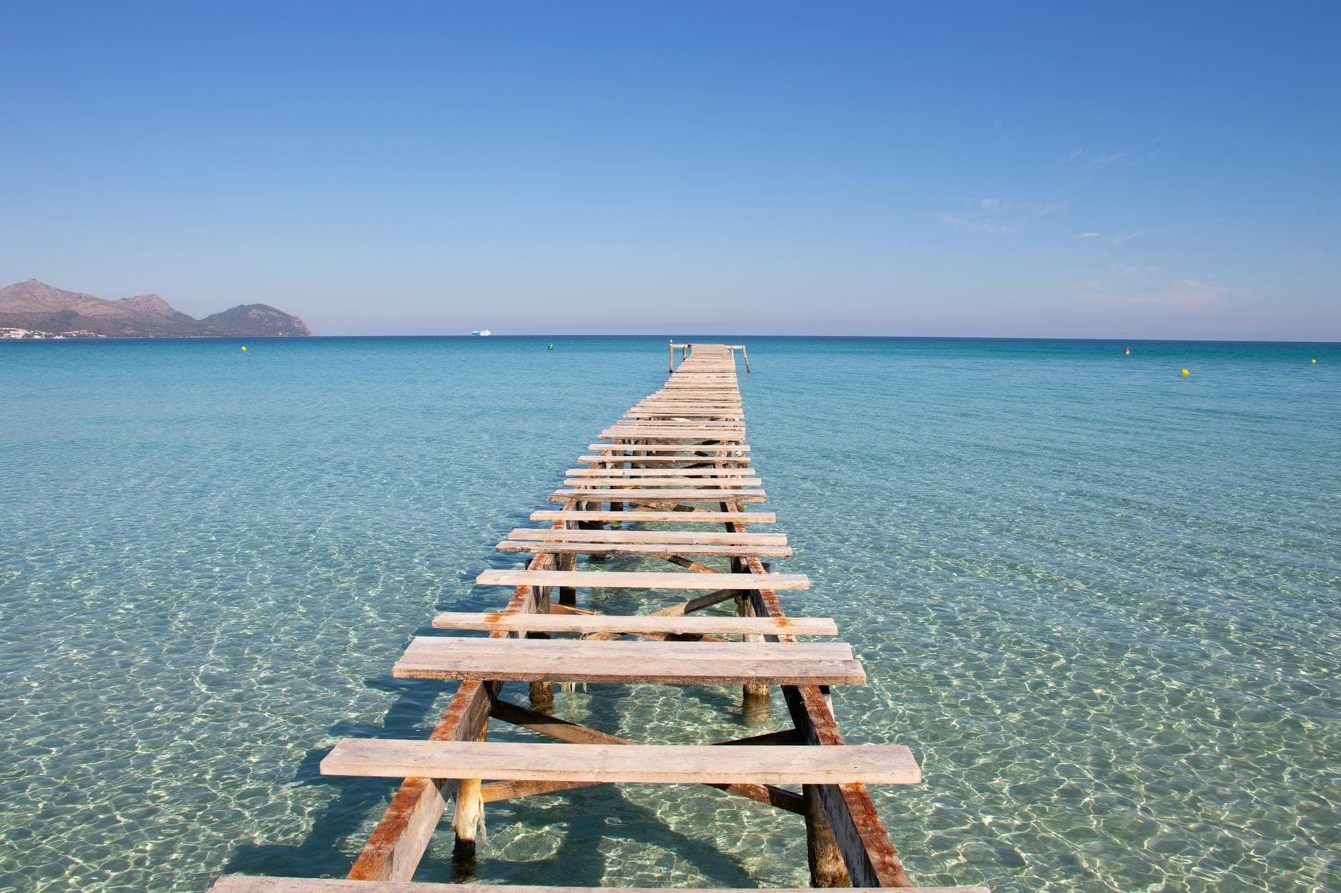Kristallklares türkisfarbenes Wasser und ein rustikaler Holzsteg, der ins Meer hinausragt, an der Playa de Muro – im Hintergrund Berge unter blauem Himmel.