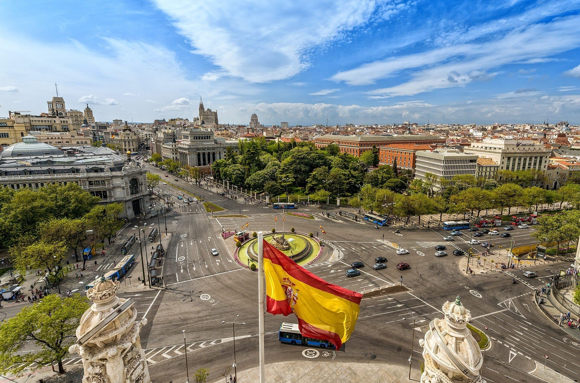 มุมมองทางอากาศของ Plaza de Cibeles ในมาดริด ซึ่งมีน้ำพุอันเป็นสัญลักษณ์ อาคารประวัติศาสตร์ ต้นไม้เขียวขจี และธงชาติสเปนโบกสะบัดใต้ท้องฟ้าสีคราม