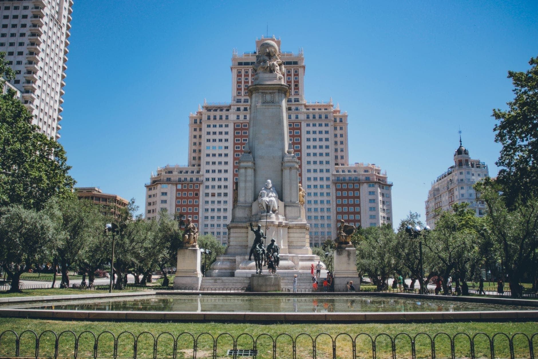 Широкий вид на Plaza de España в Мадриде: на переднем плане — величественный памятник Сервантесу, вокруг — пышная зелень и исторические здания на фоне ясного голубого неба.