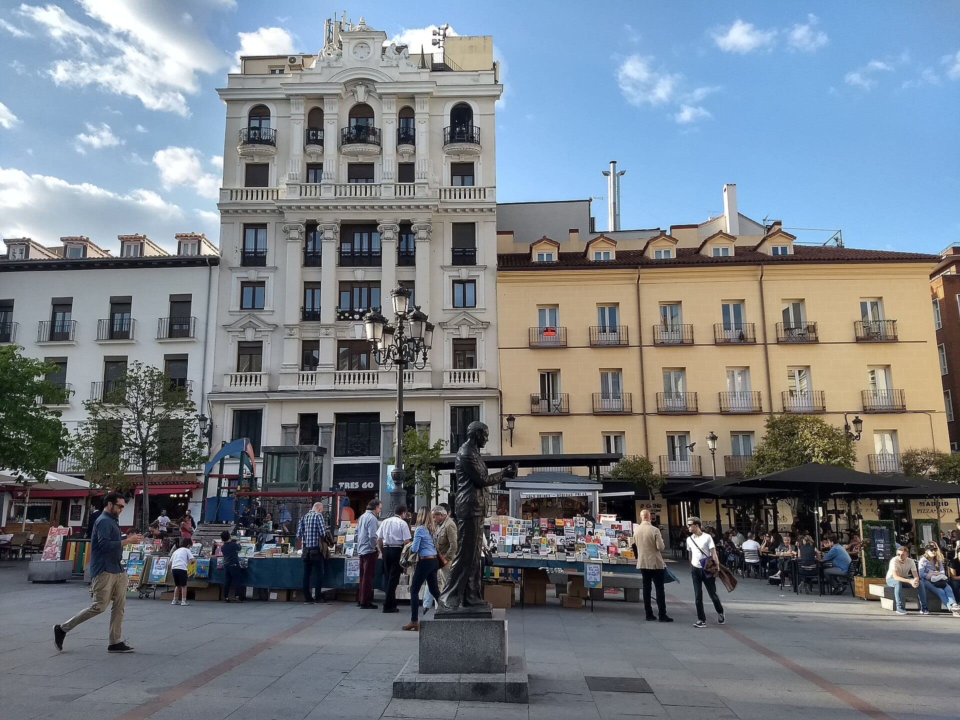 Plaza de Santa Ana ในมาดริด พร้อมอาคารประวัติศาสตร์ รูปปั้นกลางจัตุรัส แผงขายหนังสือกลางแจ้ง และผู้คนที่เพลิดเพลินกับระเบียงคาเฟ่ในวันที่แดดสดใส