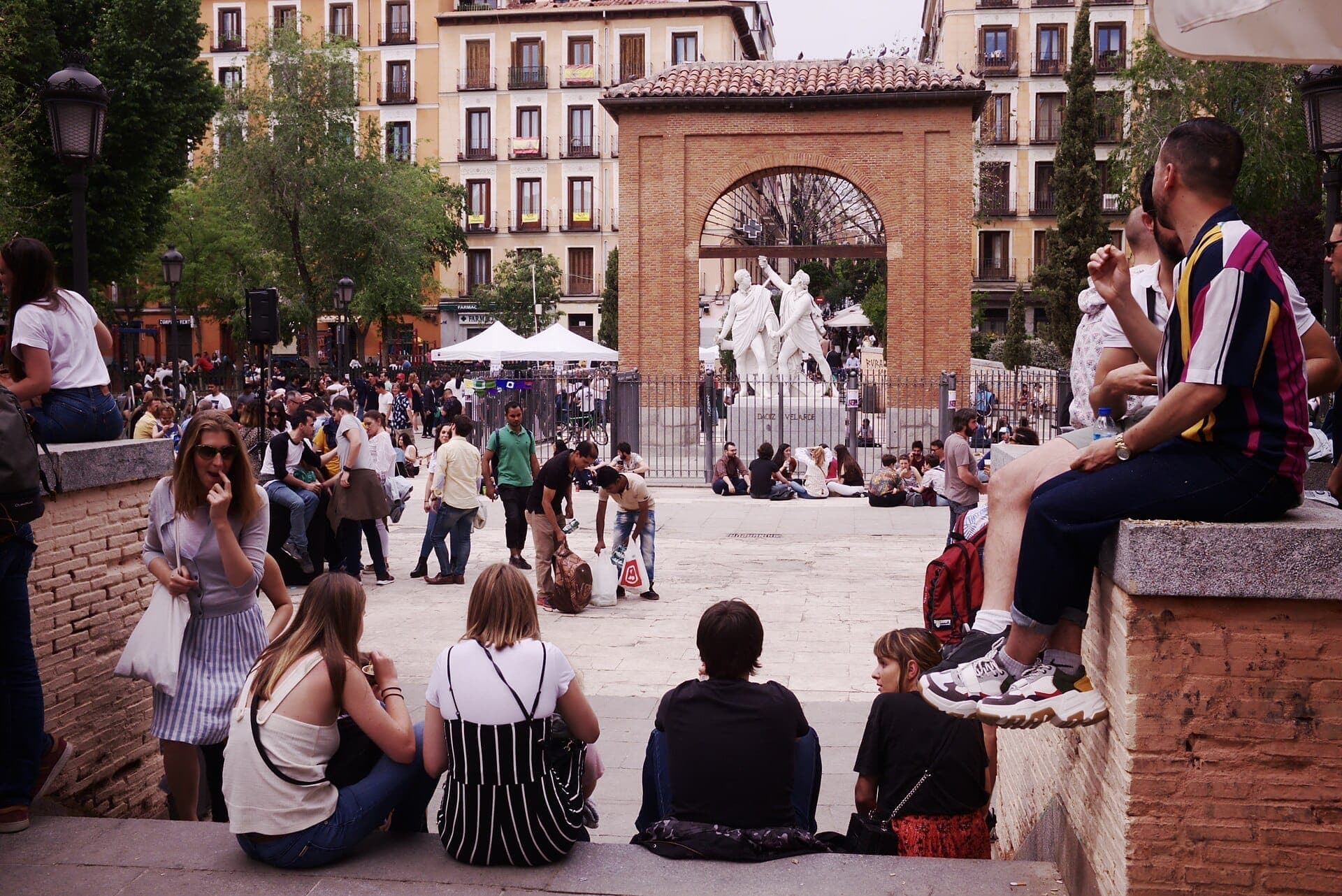 วิวของ Plaza del Dos de Mayo ในมาดริด พร้อมผู้คนที่พักผ่อนบริเวณโค้งประตูและรูปปั้นตรงกลาง ล้อมรอบด้วยอาคารอพาร์ตเมนต์หลากสีสัน