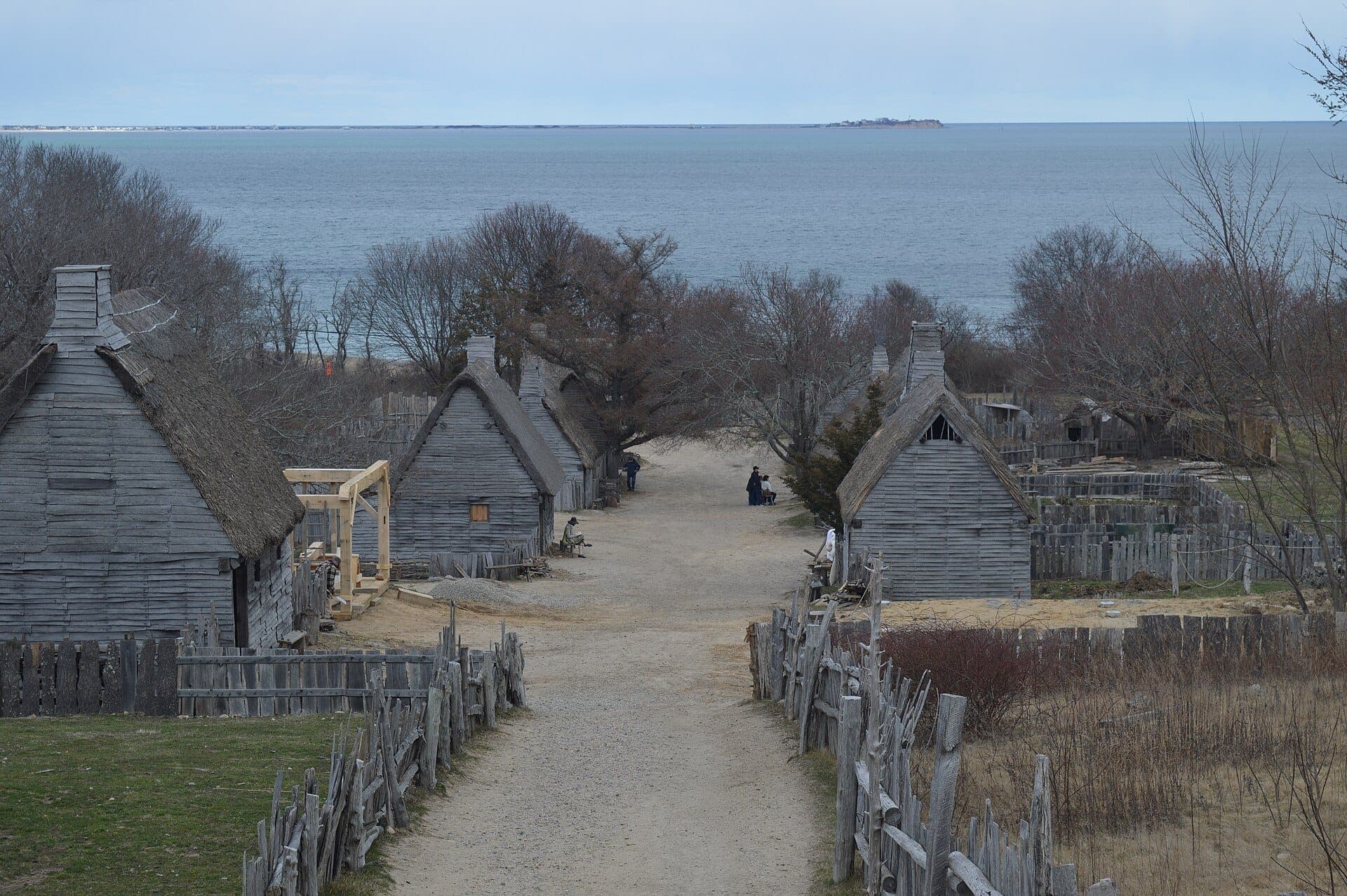 Village historique du Plimoth Patuxent Museums avec des maisons en bois vieilli, des clôtures en rondins fendus et une vue sur l'océan sous un ciel nuageux.