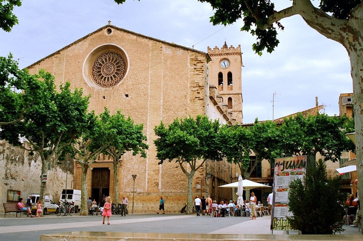 Historyczna fasada kościoła i wieża zegarowa w Starym Mieście Pollença, Mallorca, z zielonym placem, odpoczywającymi ludźmi i słoneczną atmosferą.