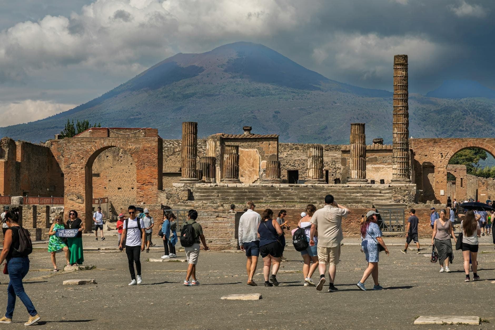 Visitantes exploram as ruínas antigas do Parque Arqueológico de Pompeia com o Monte Vesúvio imponente ao fundo sob nuvens dramáticas.