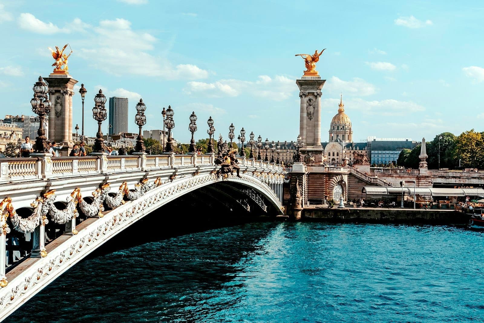 Wide view of Pont Alexandre III in sunlight, showing ornate lamp posts, gilded statues, and bright blue water with cityscape in the background.