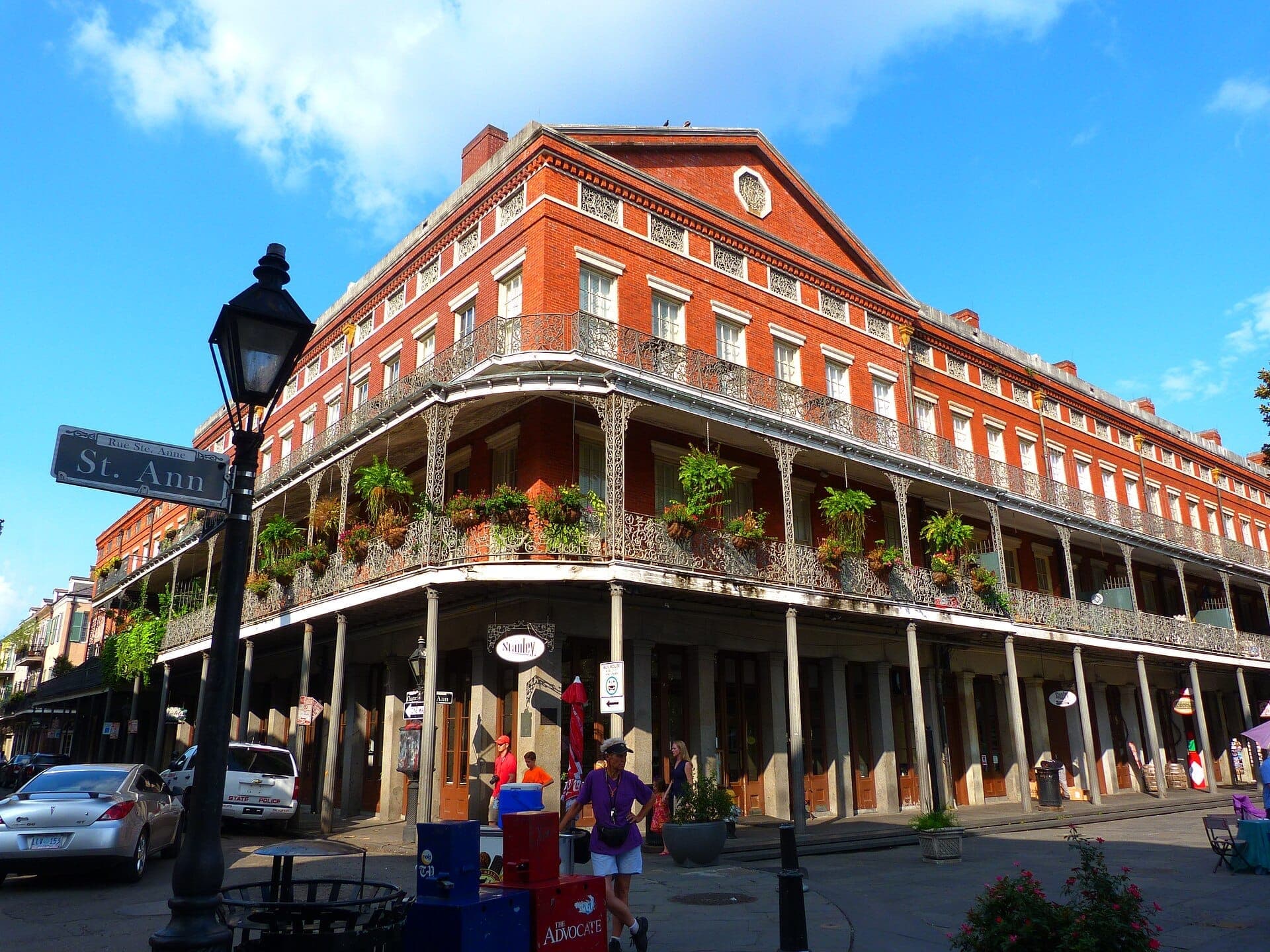 The Pontalba Buildings in New Orleans, showing red-brick facade, cast-iron balconies, street sign, parked cars, and pedestrians under a blue sky.