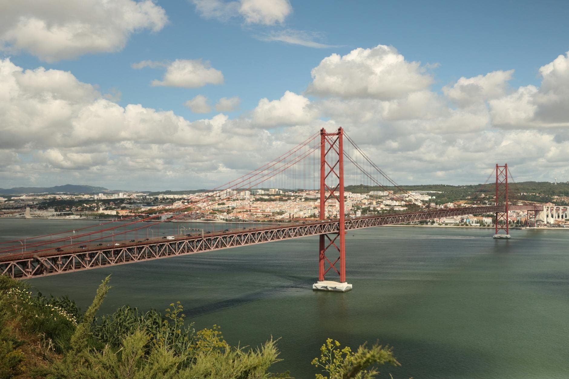 Vista panorâmica da ponte pênsil Ponte 25 de Abril cruzando o Rio Tejo, com o skyline de Lisboa e um céu azul nublado ao fundo.