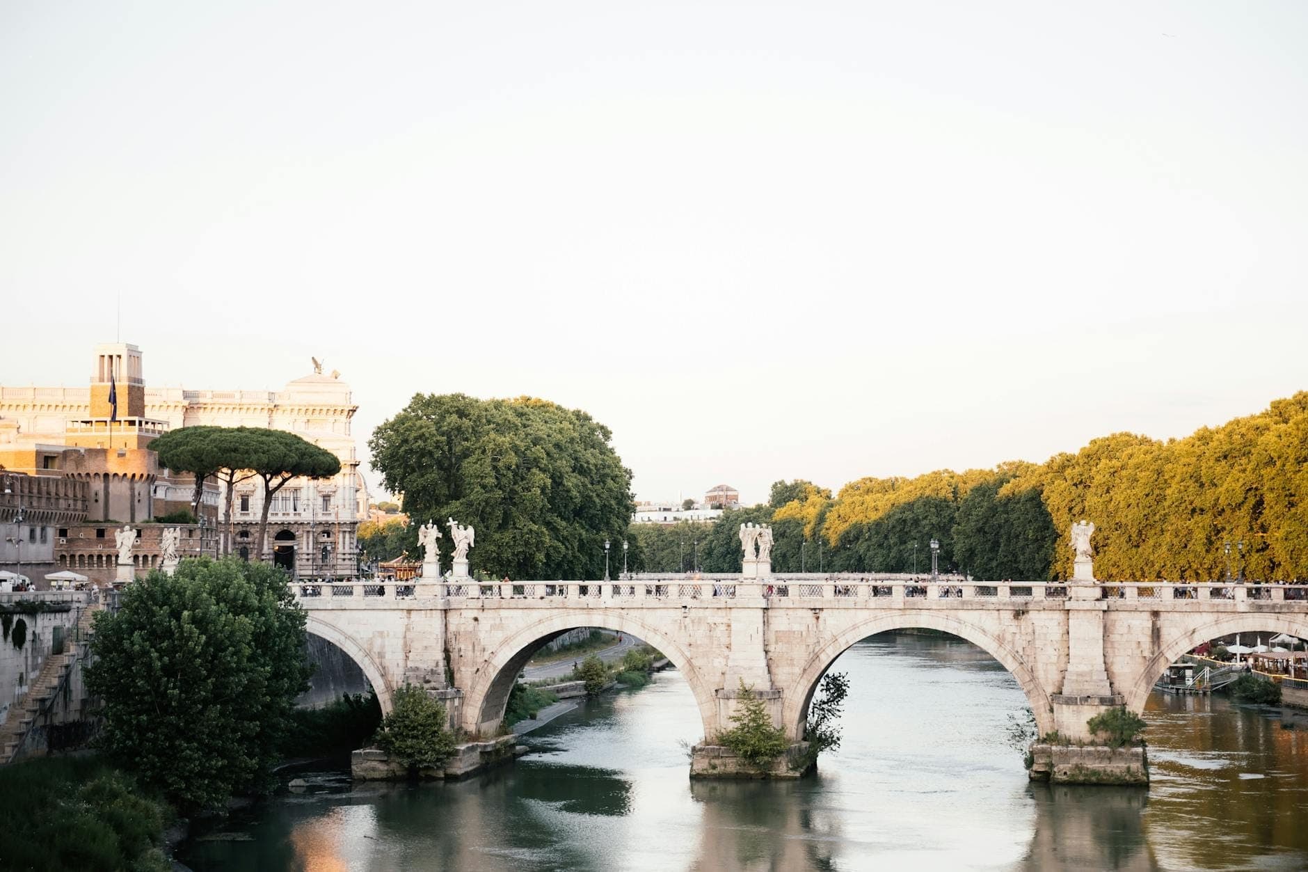 Cầu Ponte Sant'Angelo với các tượng thiên thần Baroque bắc qua sông Tiber, cây xanh tươi tốt và những công trình La Mã cổ kính dưới nắng rực rỡ.