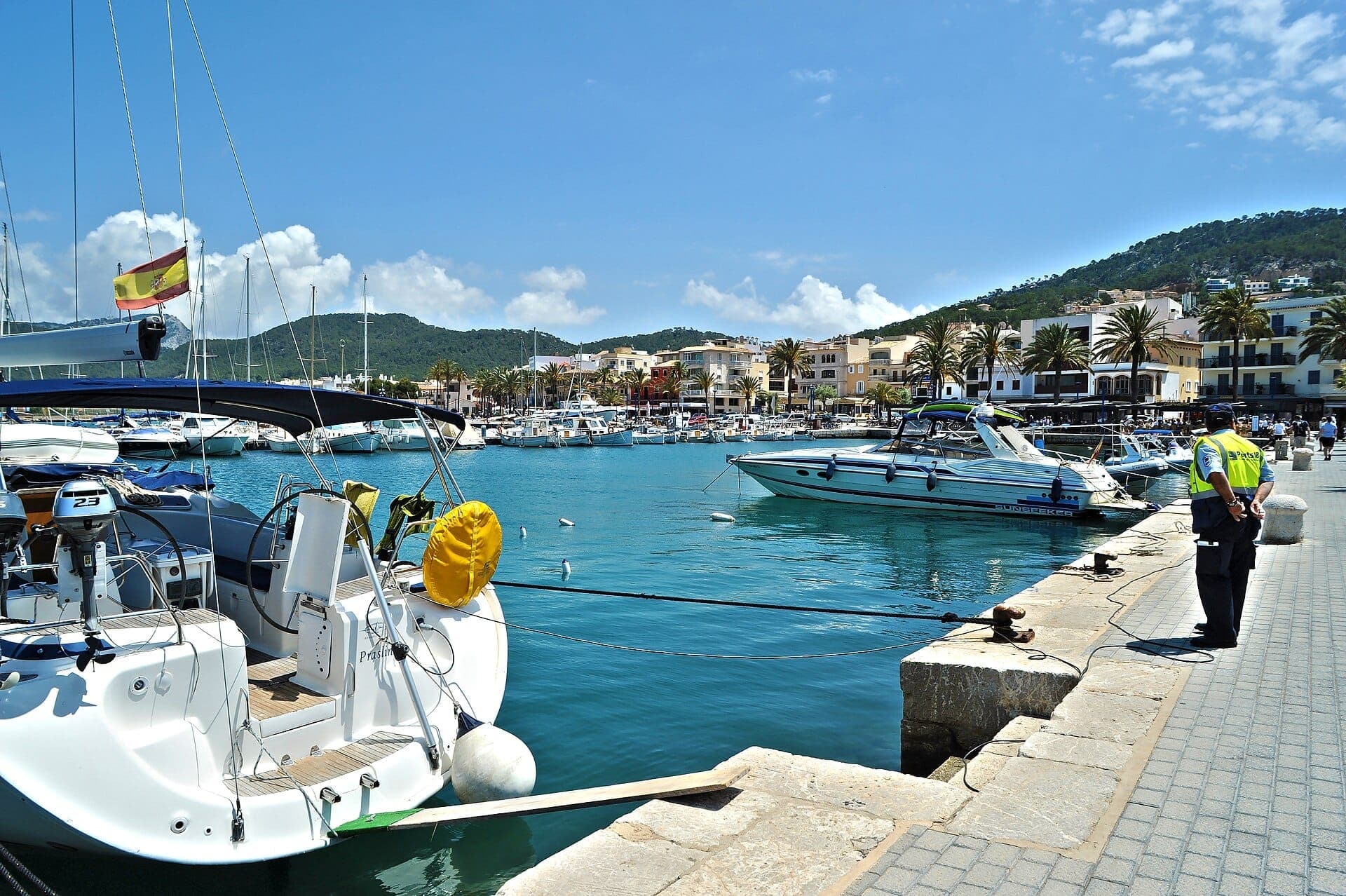 Yachten und Boote im türkisfarbenen Wasser der Marina von Port d'Andratx, gesäumt von Palmen und pastellfarbenen Gebäuden am Ufer unter einem sonnigen Himmel.
