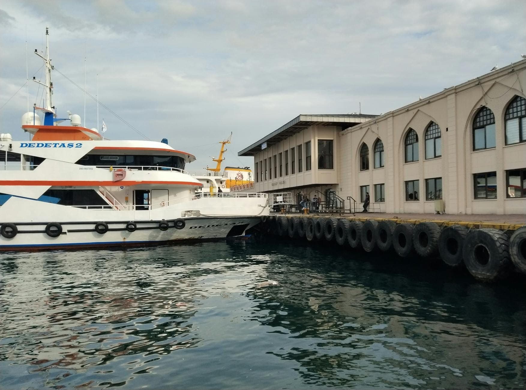 Un ferry moderno atracado en una terminal con grandes ventanales en arco, aguas tranquilas del puerto en primer plano y un cielo parcialmente nublado.