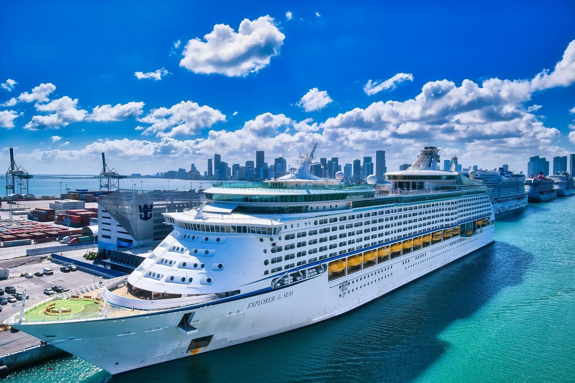 Large cruise ship docked at PortMiami terminal with downtown Miami skyline and blue skies in the background on a sunny day.
