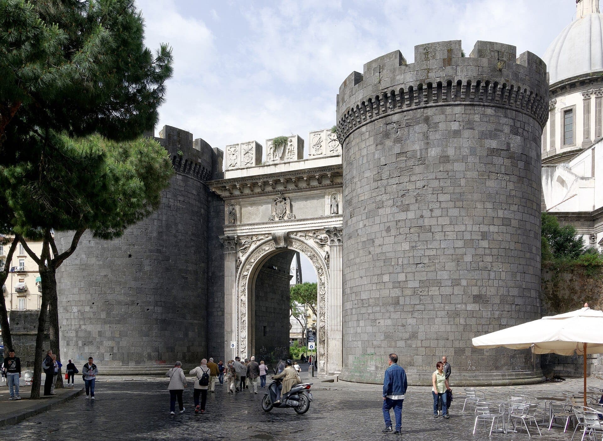 Porta Capuana em Nápoles vista de frente, com suas duas imponentes torres de mármore, o arco renascentista e algumas pessoas caminhando ao redor.