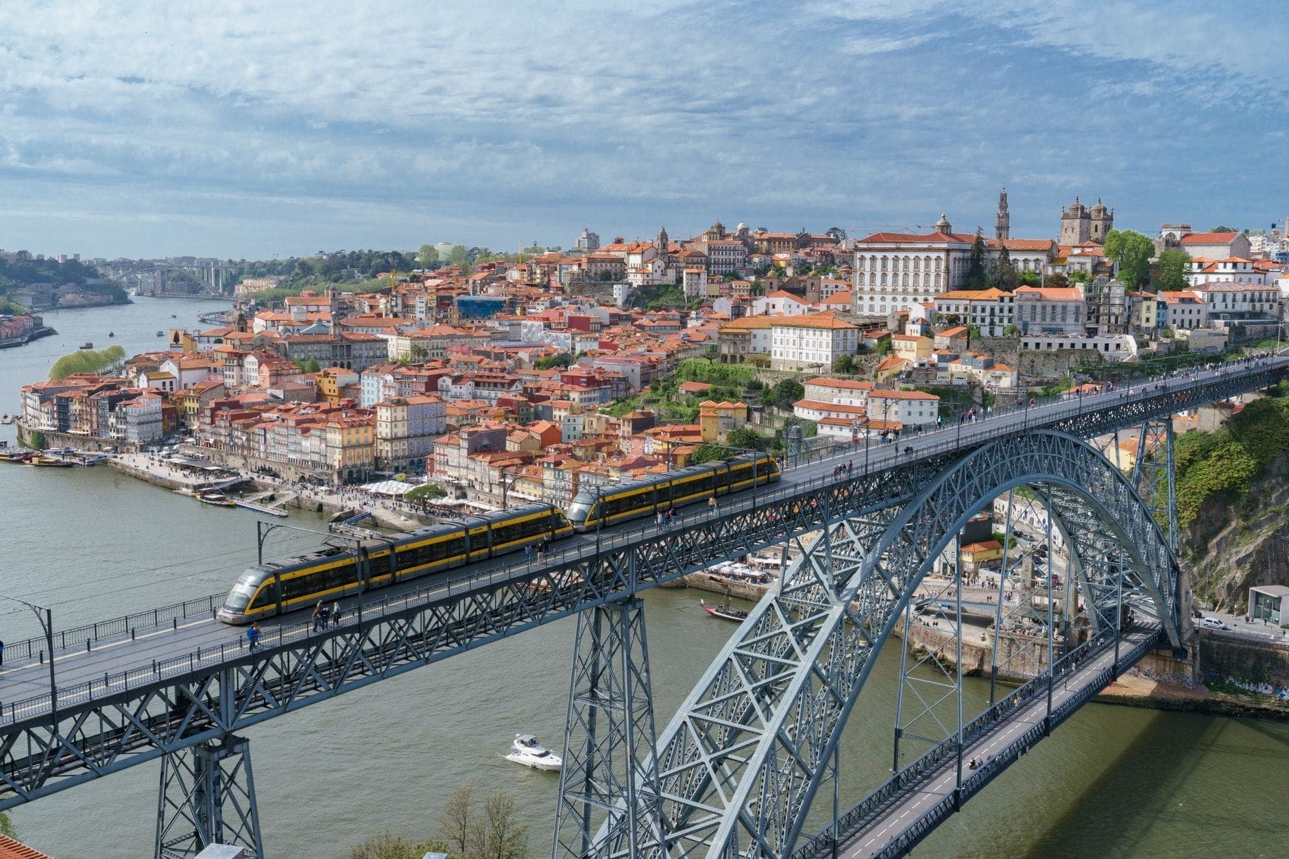 Panoramablick auf Porto mit der ikonischen Dom-Luís-I-Brücke, einer Metro-Überquerung und der historischen Stadtsilhouette am Douro-Ufer unter strahlend blauem Himmel.