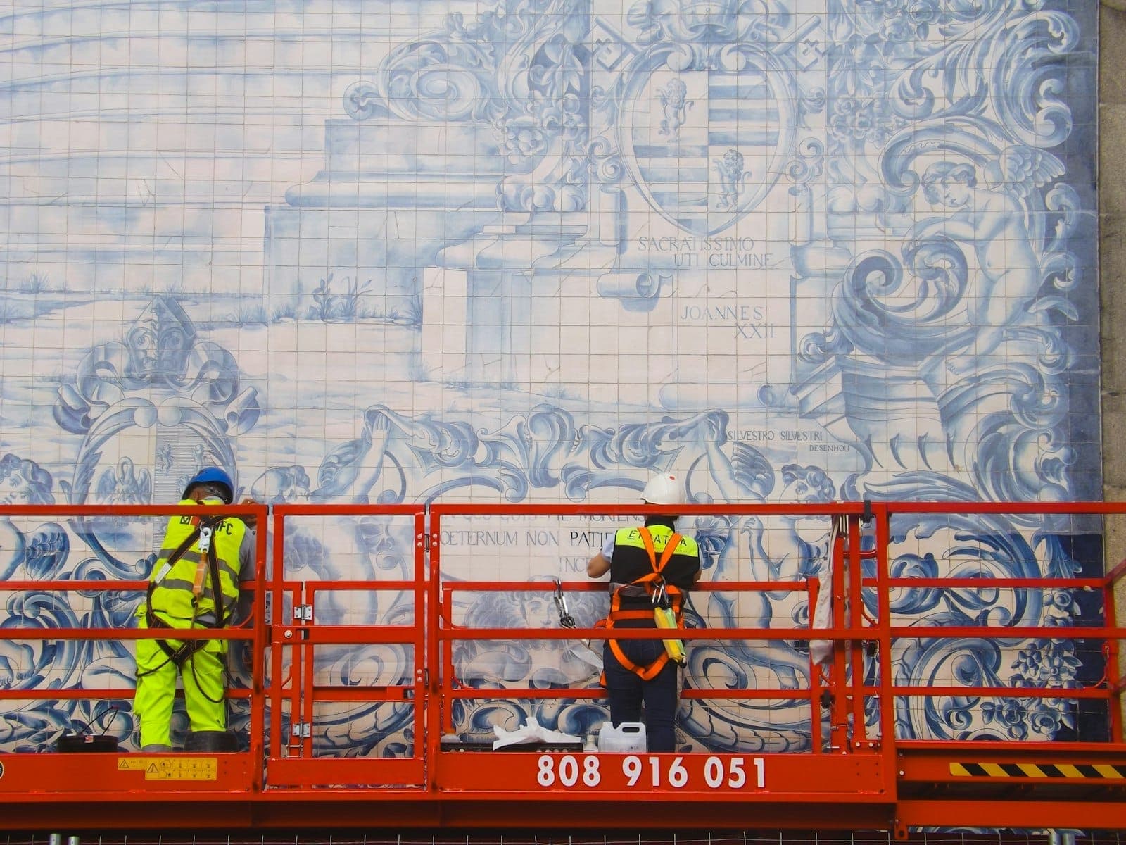 Trabalhadores em uma plataforma elevatória restaurando um grande mural de azulejos azuis e brancos na fachada de um edifício no Porto, Portugal.