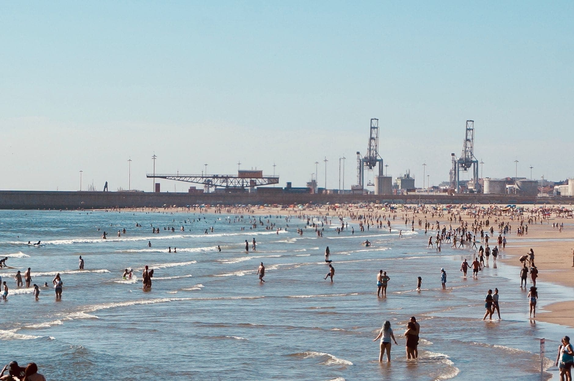 Belebter Stadtstrand mit schwimmenden und watenden Menschen im Flachwasser, Kräne und Hafenstrukturen im Hintergrund.