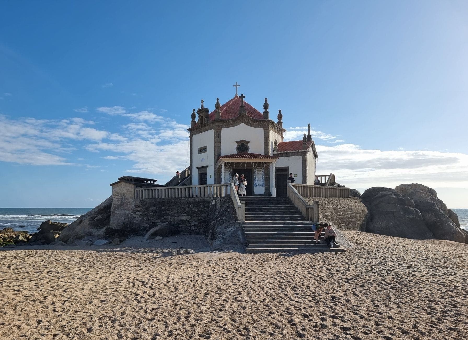 Kapelle am Strand von Vila Nova de Gaia unter blauem Himmel, mit Sand, Felsen und dem Atlantischen Ozean im Hintergrund.