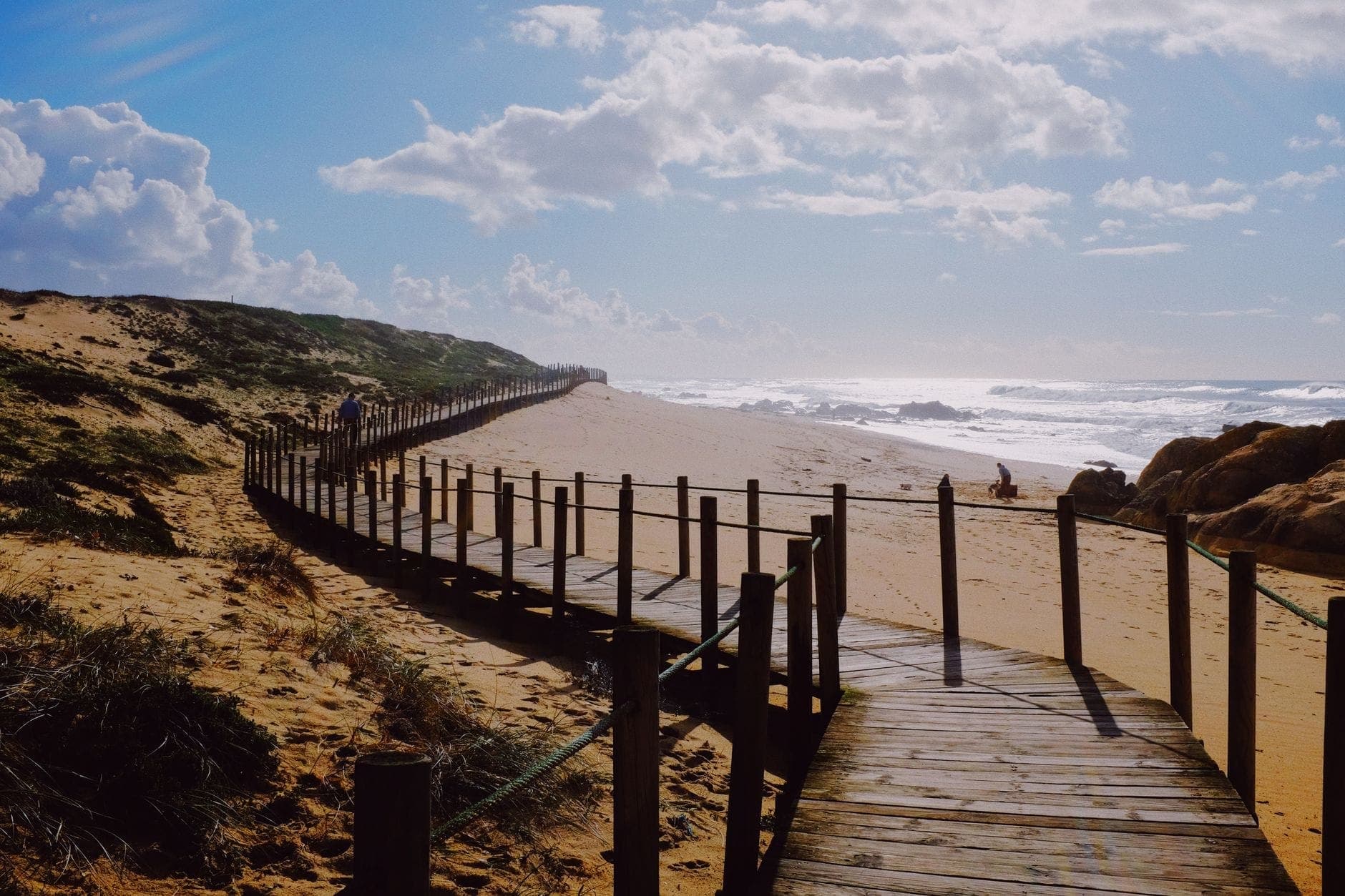 Ein Holzsteg schlängelt sich an einem Sandstrand bei Porto entlang, mit sanften Wellen, blauem Himmel und Menschen, die in der Ferne die Küste genießen.