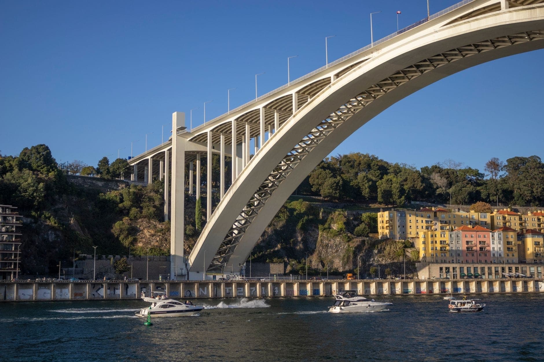 Vue panoramique du Pont de l'Arrábida enjambant le fleuve Douro à Porto, avec des bateaux et des bâtiments colorés au bord de l'eau sous un ciel bleu dégagé.