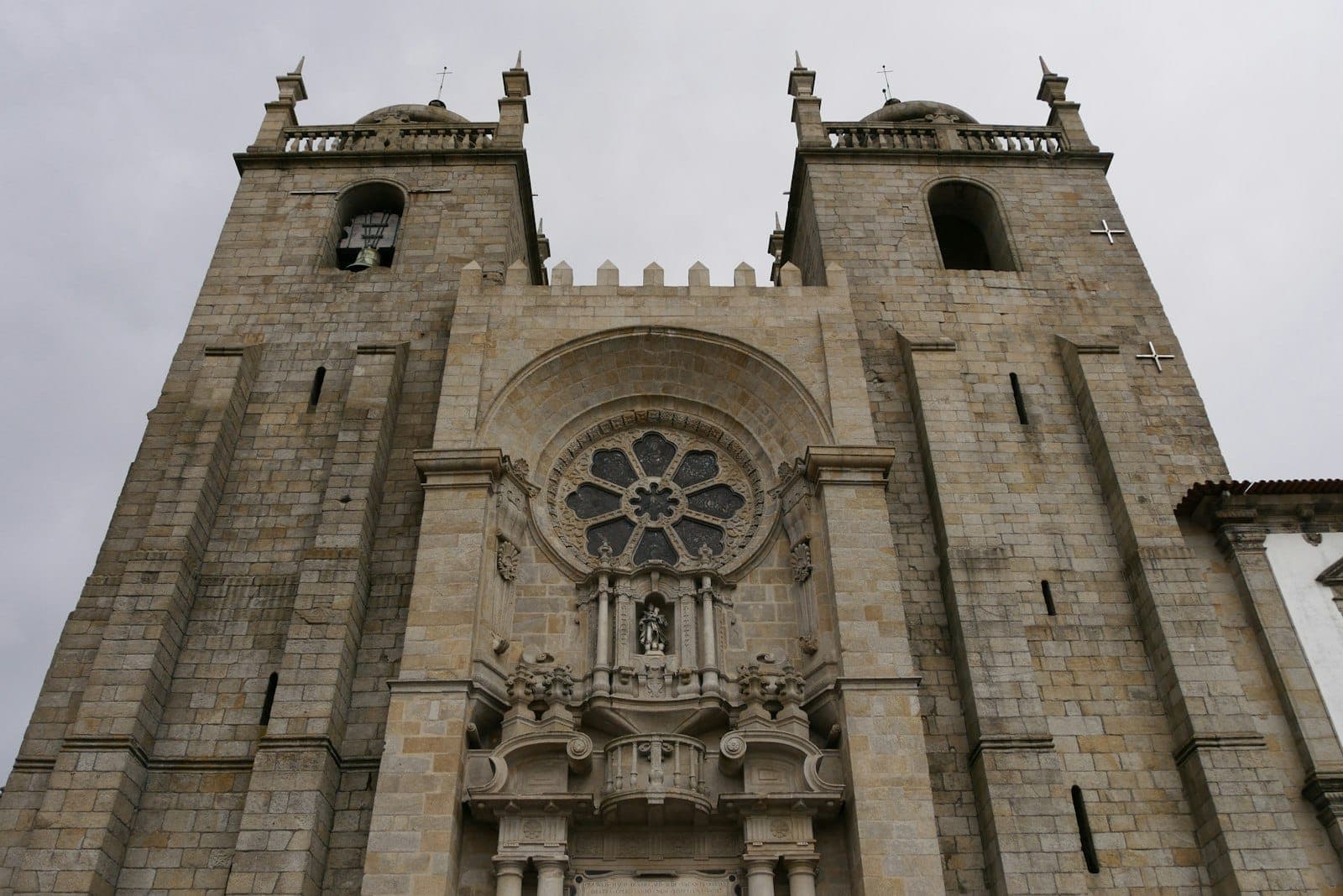 Die imposante Steinfassade der Kathedrale von Porto mit Doppeltürmen, Bogeneingang und kunstvollem Rosettenfenster unter bewölktem Himmel.
