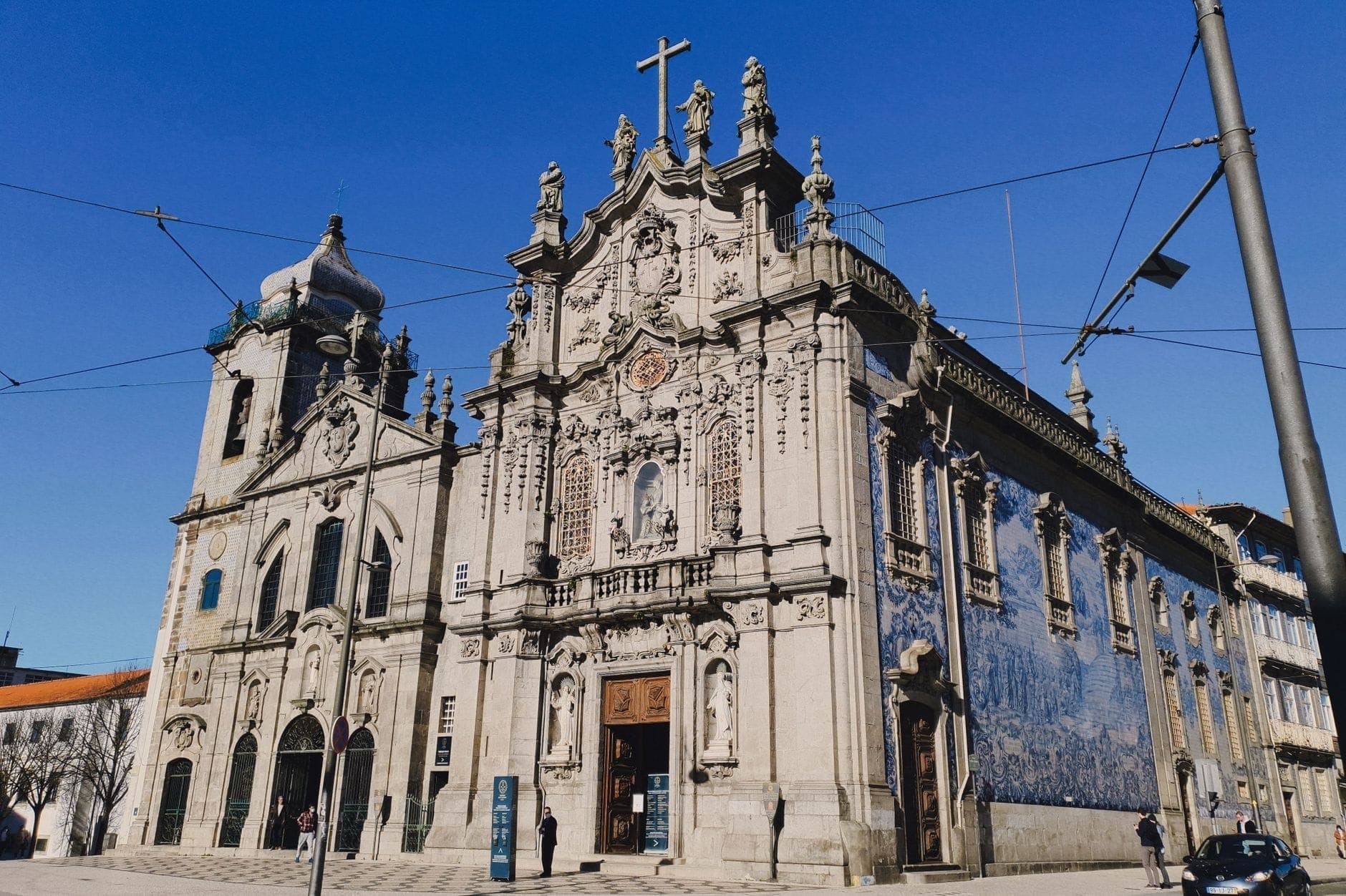 Barocke Kirchenfassade in Porto mit aufwändigem Steinmetzwerk, Statuen und charakteristischen blauen Azulejo-Kacheln unter strahlend blauem Himmel. Straße und vereinzelte Menschen im Vordergrund.