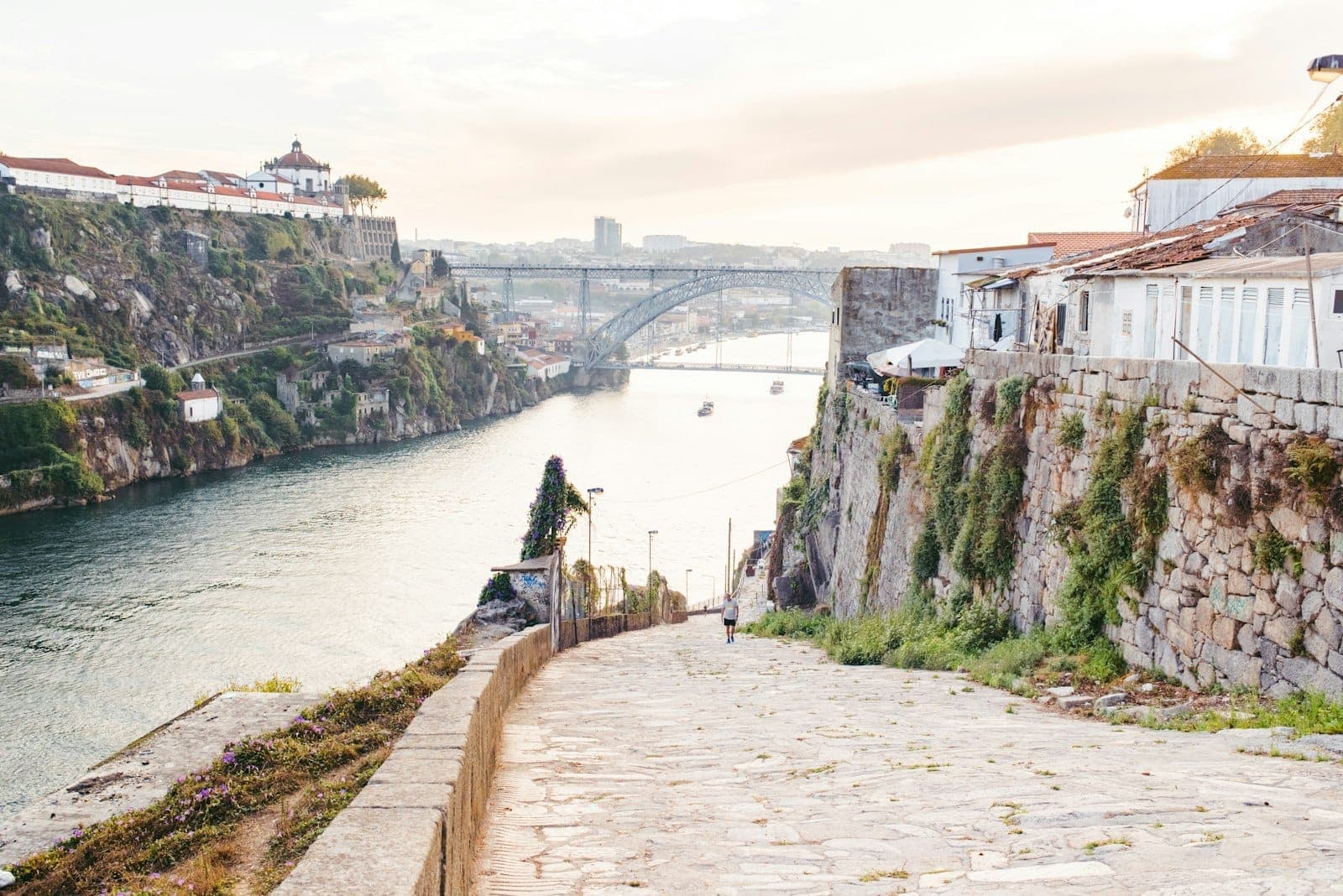 Un tranquillo vicolo in pietra scende verso il fiume Douro, con lo storico ponte in ferro di Porto sullo sfondo e gli edifici illuminati dal sole su entrambe le rive.