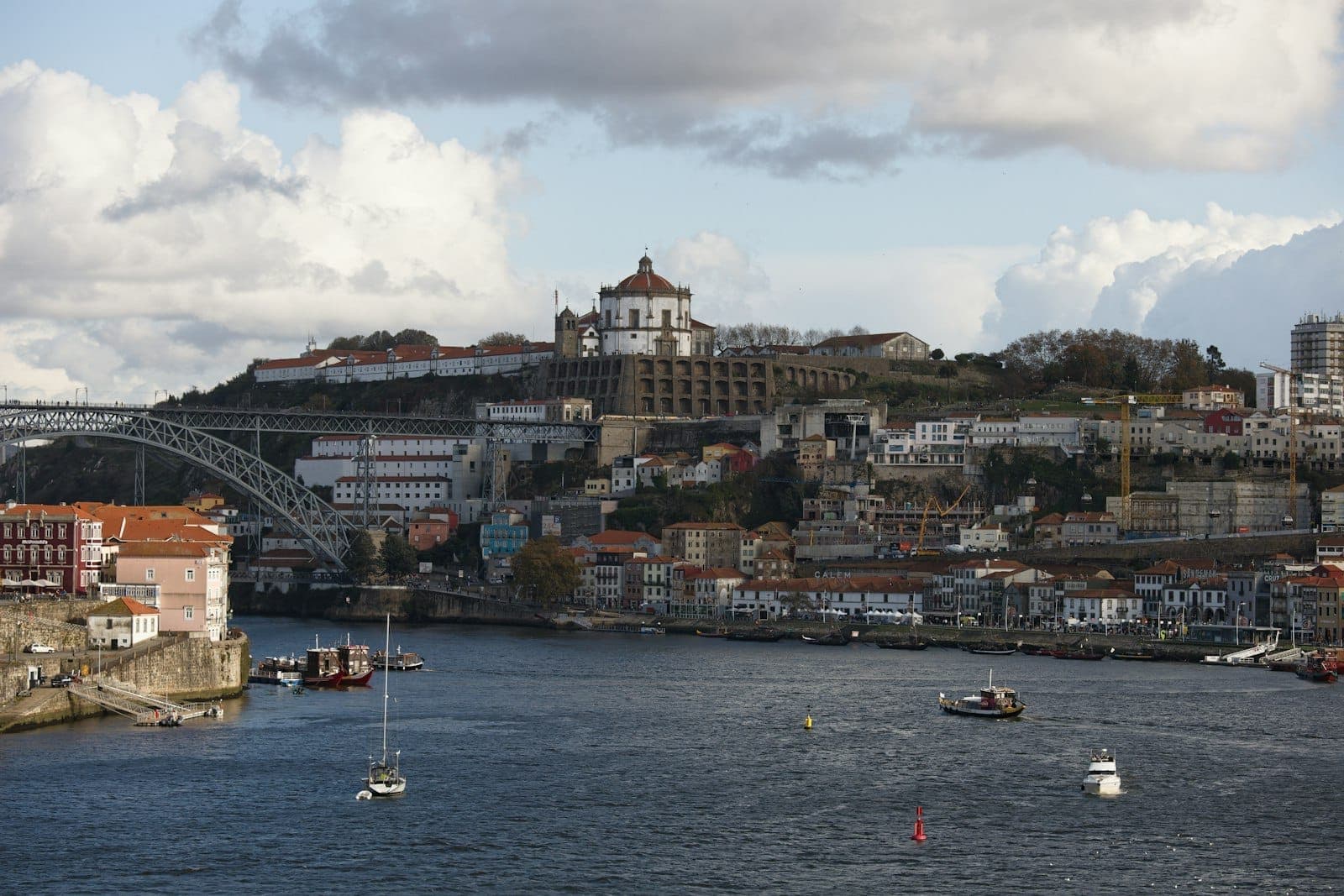 Vista del río Duero hacia Vila Nova de Gaia con bodegas de vino de Oporto, edificios coloridos, el icónico Puente Dom Luís I y el Monasterio de Serra do Pilar en la colina.