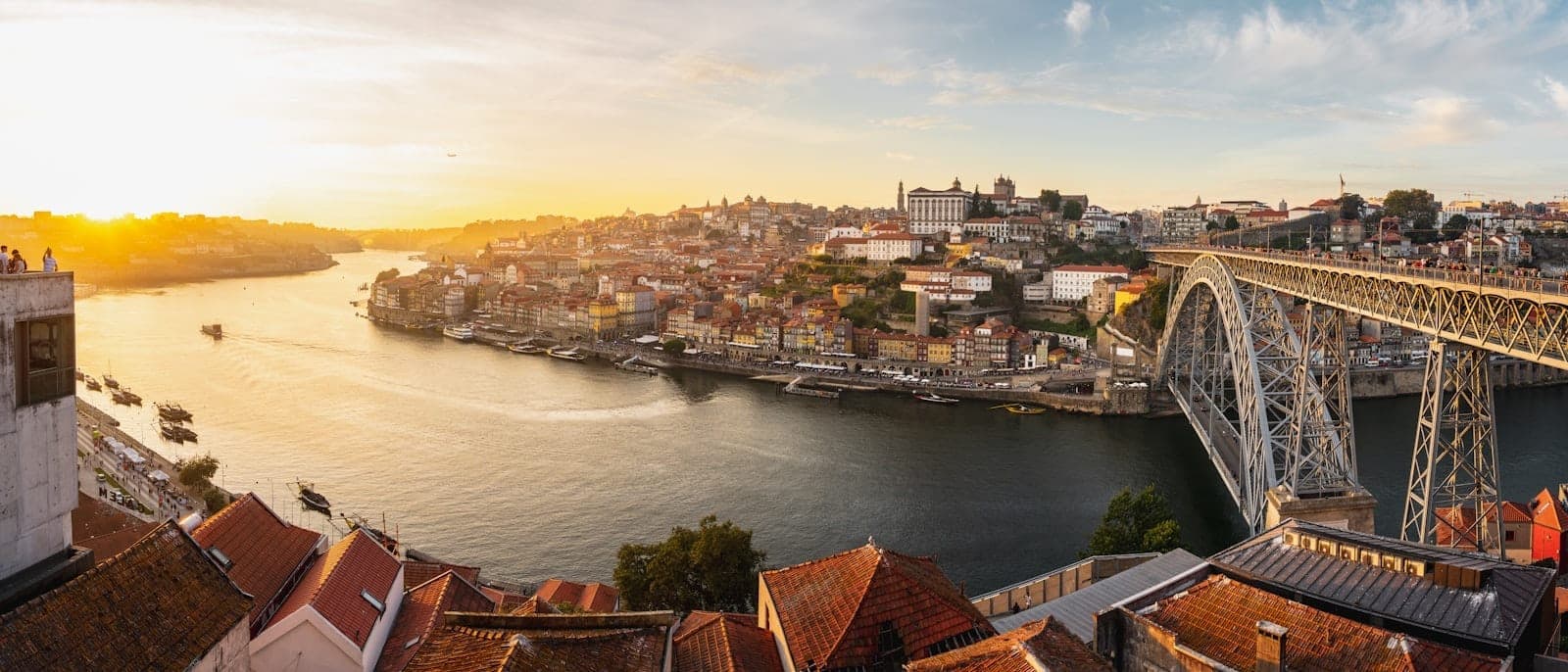 Vista panoramica al tramonto sul lungofiume storico di Porto e il Ponte Dom Luís I, con i tetti color terracotta e i monumenti della città che si affacciano sul fiume Douro.