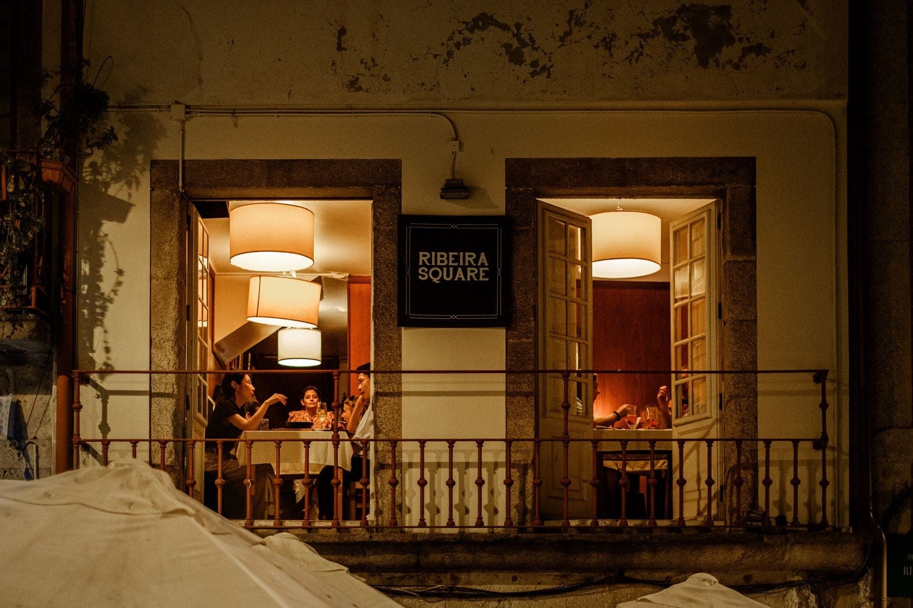 Vista con luce calda di un ristorante in Piazza Ribeira a Porto di notte, con i commensali visibili attraverso le finestre aperte.